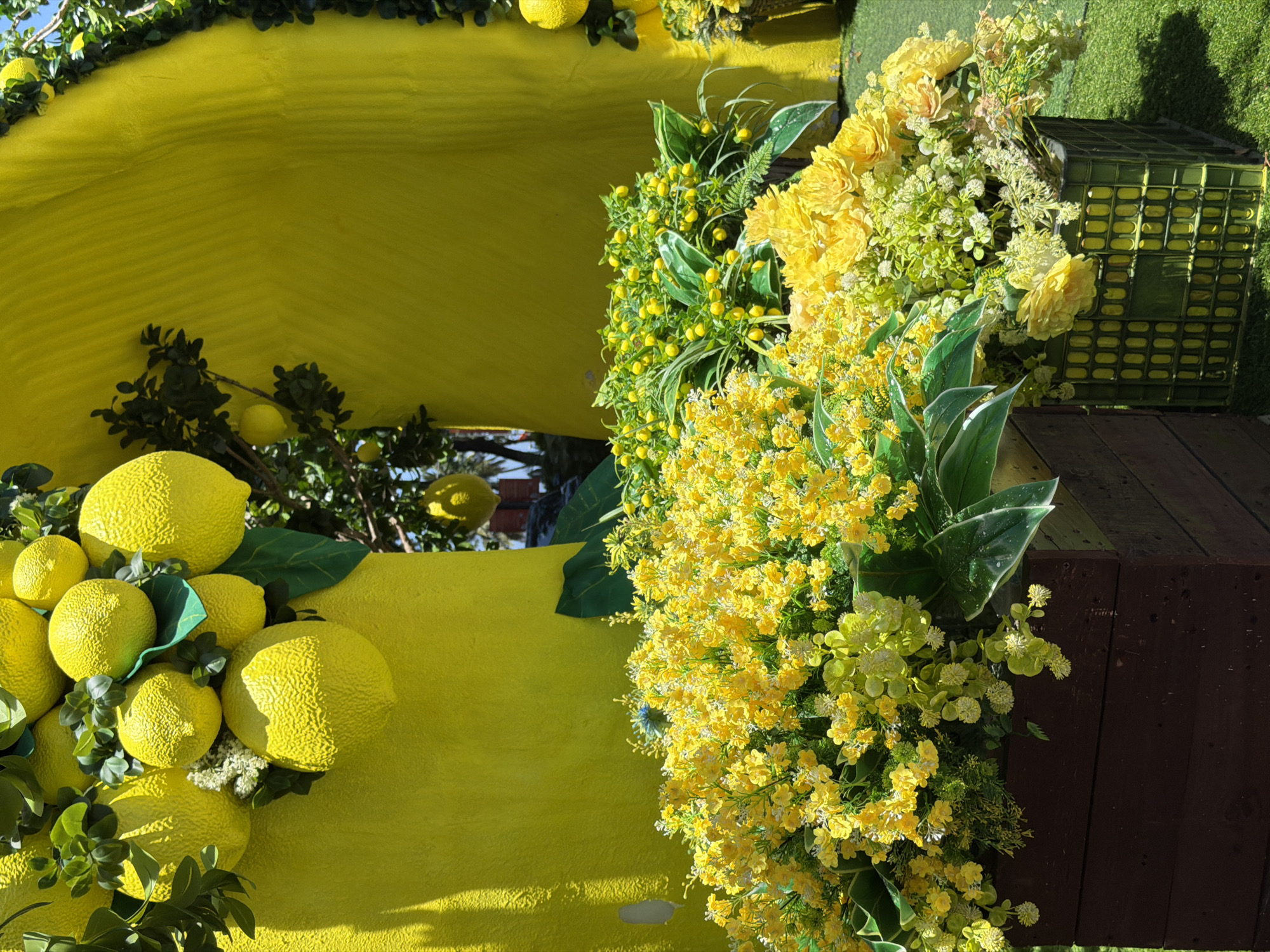 The image shows a brightly decorated structure with yellow lemons and yellow flowers. A yellow, possibly painted, surface serves as a backdrop, adorned with lemons and foliage. To the right is an abundant arrangement of yellow flowers, varying in size and texture. A dark brown, wooden surface partially covers the right side of the frame, and an olive green crate sits near the top. The image is taken from a close perspective, emphasizing the details of the decorative elements. The location is Nha Trang Ward, Vietnam.