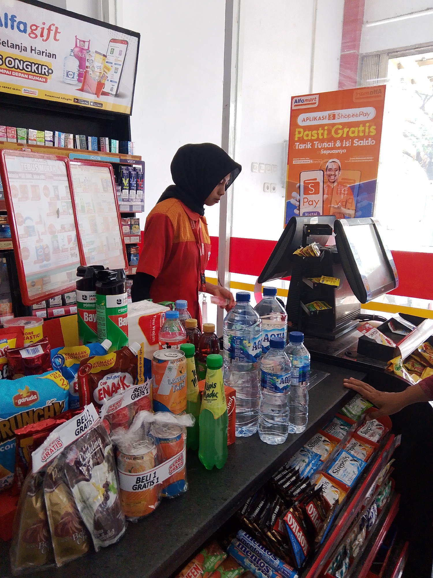 The image depicts the interior of an Alfamart convenience store, specifically at the checkout counter in Bolo Krajan, Indonesia. A female cashier, wearing an orange and red uniform and a black hijab, is standing behind the counter. She is looking down at the items being processed. The counter is cluttered with various products, including bottled water, canned drinks, snacks, and toiletries. A cash register with two screens is prominently displayed. A promotional poster for "ShopeePay" is visible in the background, advertising "Pasti Gratis" (Certainly Free) offers. The environment appears to be typical of a convenience store, with shelves stocked with merchandise and promotional displays. The lighting suggests an indoor setting, possibly with natural light entering from an unseen source.