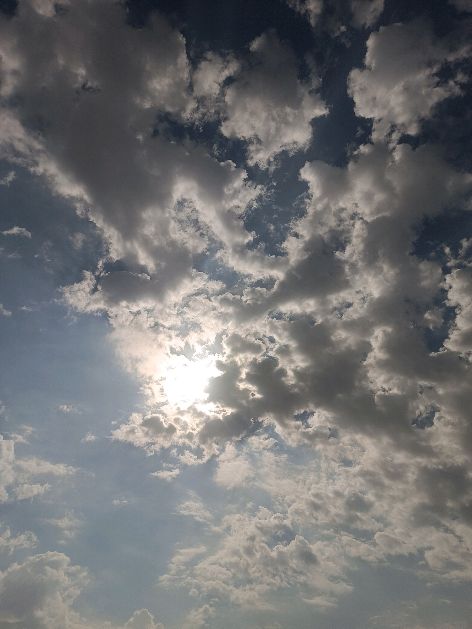 The image shows a sky filled with a mixture of dark gray and white clouds, with patches of blue sky visible. The sun is partially obscured by a large cluster of clouds in the lower right quadrant of the image, casting a bright, hazy glow. The clouds are dense and appear to be moving across the sky, creating a dynamic and somewhat dramatic scene. There are no people, buildings, or any other discernible objects or features in the image, as it is solely focused on the sky. The environment depicted is an outdoor sky scene, not specific to any particular location in Myrza-Ake, Kyrgyzstan, or any other city.