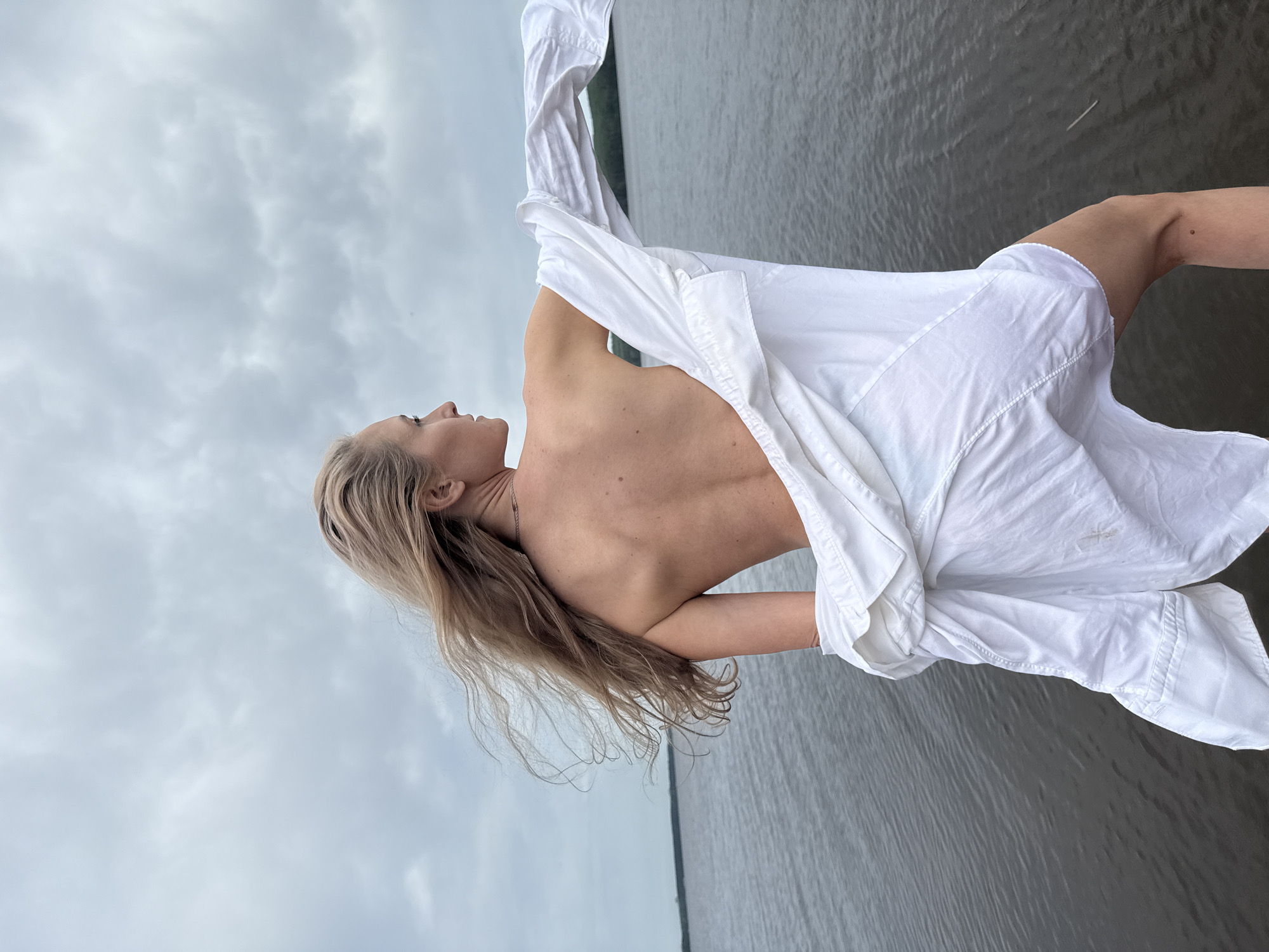 A woman with long blonde hair is standing near a body of water under a cloudy sky. She is partially dressed in a white shirt. The setting appears to be near a shoreline, based on the water's edge in the background. The location is the city of Chaikovsky, Russia.