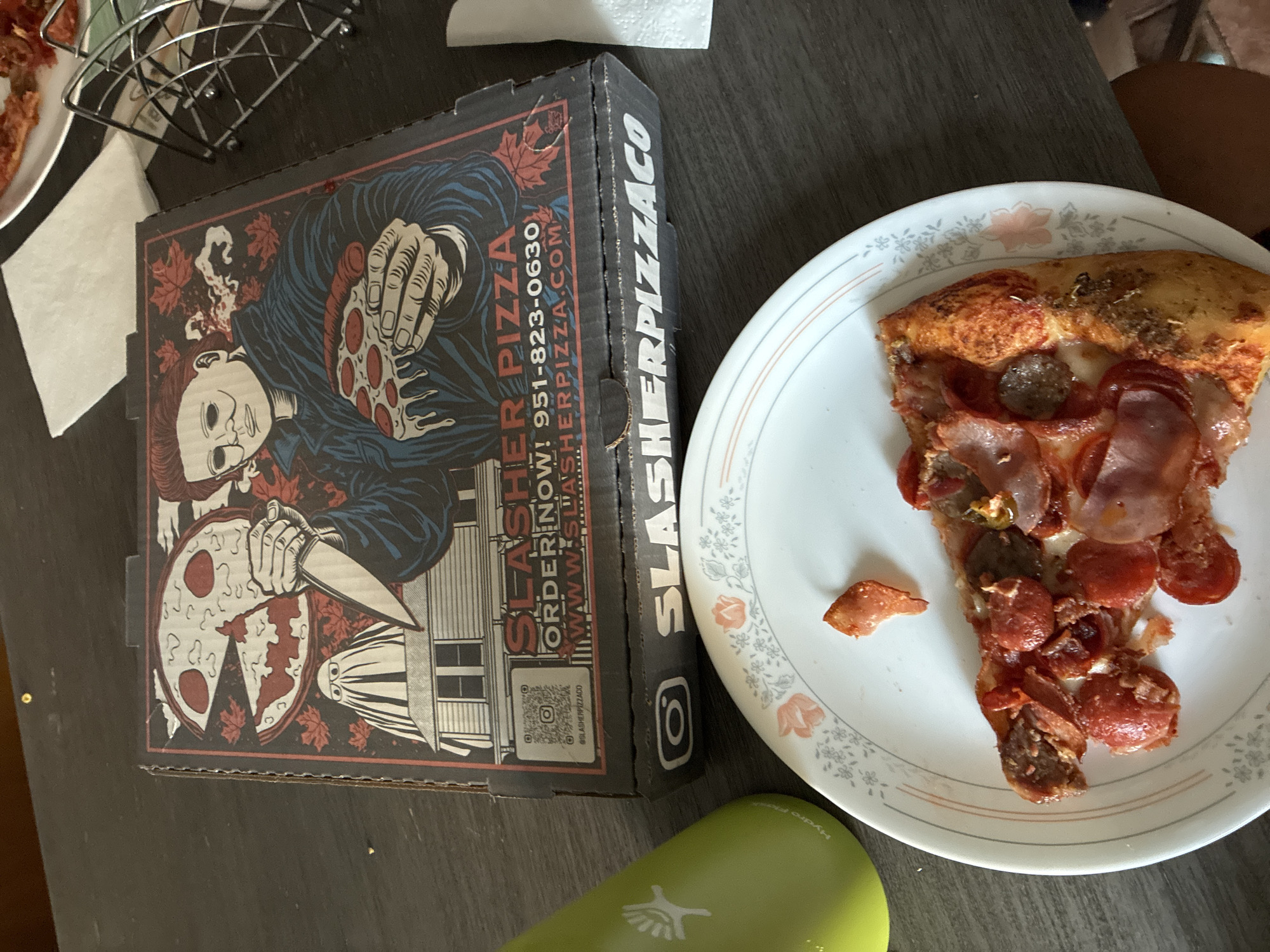 A pizza box is on a dark wooden table, to the left is a green Hydro Flask. In the foreground is a white plate with a slice of pepperoni and bacon pizza. The pizza box has an illustration of Michael Myers from the movie Halloween holding a knife and a slice of pizza. The box also displays the text "SLASHER PIZZA" and a phone number "951-823-0630" and the website "WWW.SLASHERPIZZA.COM". There is also an Instagram logo and the handle "@SLASHERPIZZACO" visible on the box. The scene is indoors, likely a home, given the table and the pizza. The location of East Hemet, United States of America is not ascertainable from the provided image.