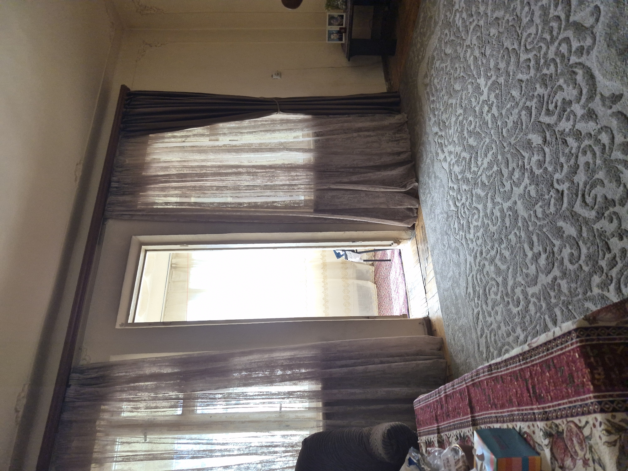 This image, taken in Yerevan, Armenia, captures an indoor room, likely a living area, bathed in natural daylight. The walls and ceiling are light-colored, showing signs of wear and minor discoloration, including what appears to be mold or water damage near the ceiling and along doorframes.
A prominent feature is a large opening, either a wide window or an open doorway, on the left side of the room. Bright sunlight streams through sheer, light-colored inner curtains. These are framed by heavier, dark brown outer curtains, one section of which is tied back. Beyond this opening, another room or exterior space is partially visible, showing a patterned light-colored curtain and a glimpse of a red-patterned rug.
The floor on the right side of the room is covered by a large, textured grey rug with an intricate, swirling damask-like pattern. In the background, a dark console table holds a few small framed pictures. In the foreground, a dark, upholstered armchair or sofa is visible, positioned next to a low table. This table is draped with a richly patterned tablecloth featuring a red and white border and a main design incorporating pomegranates and floral motifs. Several items, including plastic packaging and a small, colorful box, are placed on the table. What appears to be steps or stairs are visible in the bottom-left of some cropped views, adjacent to the patterned rug.
The bright light suggests it is daytime, likely a sunny morning or afternoon. The traditional patterns on the rug and the pomegranate motif on the tablecloth provide subtle cultural cues consistent with the provided location context of Armenia. No people are visible, and there is no discernible text in the image. The scene depicts a lived-in, domestic setting with elements of traditional decor.