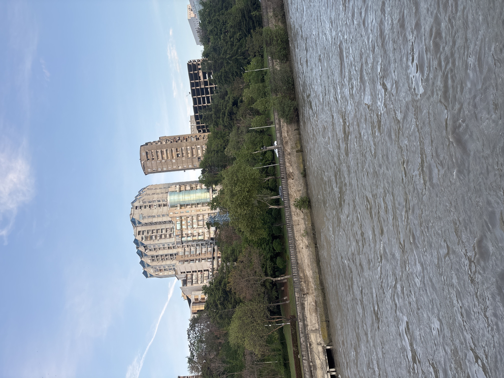 The image shows a view in Tbilisi, Georgia. A muddy river flows along a concrete embankment lined with trees and a park. Adjacent to the park are several high-rise apartment buildings, one of which is unfinished and skeletal. The sky is light blue with some contrails. No people are visible in the frame.