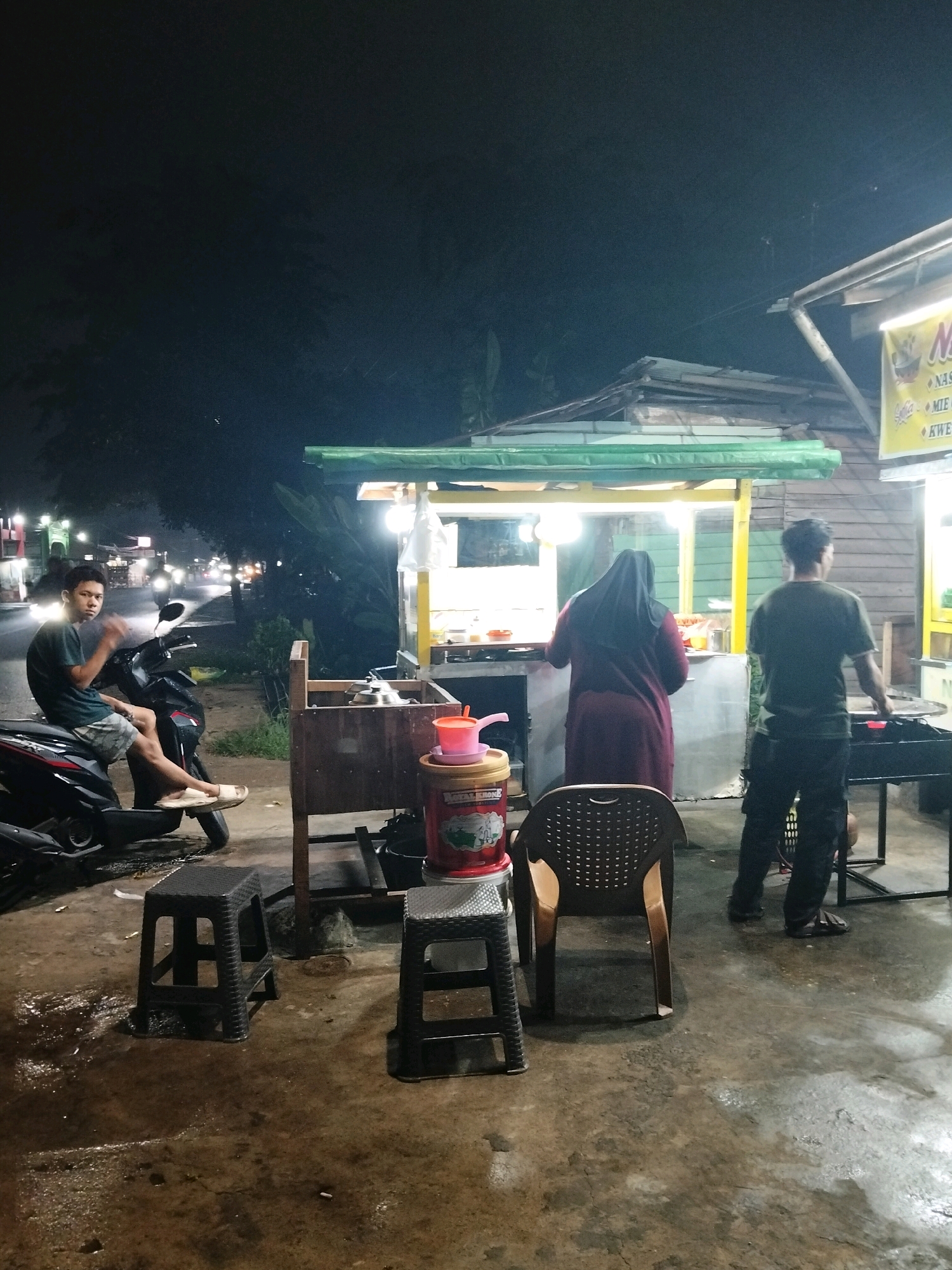 The image depicts a street-side food stall at night in Arang Limbung, Indonesia. A young man in a green t-shirt and checkered shorts sits on a black scooter on the left. To the right of the scooter, a food stall is set up with a wooden counter. A woman wearing a dark hijab and a maroon outfit is seen standing behind the stall, facing away from the camera. Another person, a man in a green t-shirt and dark pants, stands to the right of the stall, also facing away from the camera, attending to a grill. Several plastic stools are placed on the wet concrete ground in front of the stall. A yellow banner with Indonesian text, likely advertising menu items, hangs on the stall. The street behind the stall is illuminated by streetlights and the headlights of passing vehicles, indicating it is an active urban area. The overall scene is consistent with a typical night market or roadside eatery.
