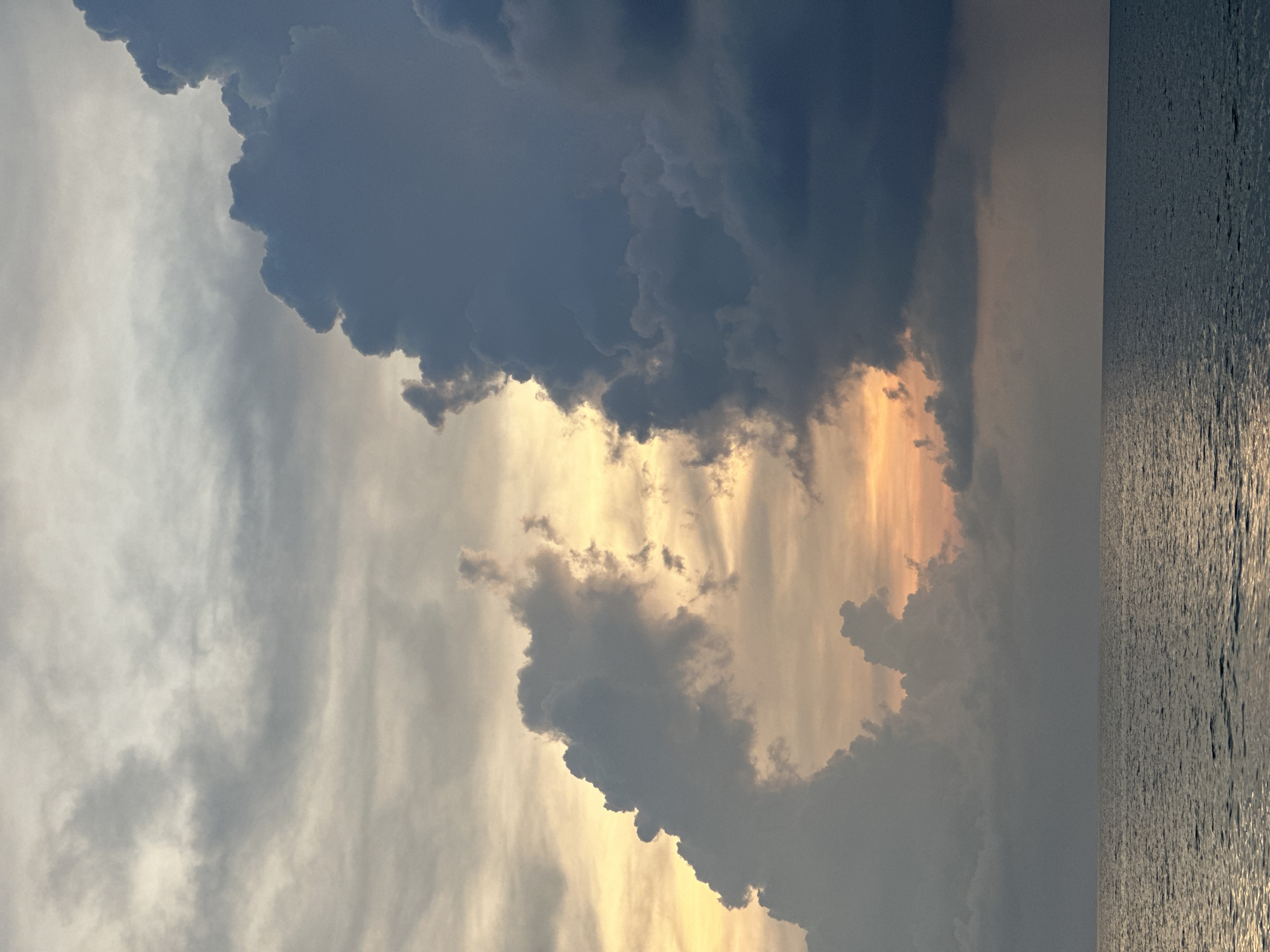 The image shows a view over water towards a dramatic sky with storm clouds and golden light. The location appears to be Choeng Thale, Thailand, as the photograph's attributes indicate this place.
The sky is divided between dark, heavy storm clouds in the upper portion, giving way to a space with bright, golden light and lighter clouds closer to the horizon. The clouds have a turbulent, textured appearance. To the right, the water surface reflects a faint glow, suggesting sunlight. The water is still, with slight ripples causing glittering light. The horizon line is relatively straight. There is no indication of human presence.
