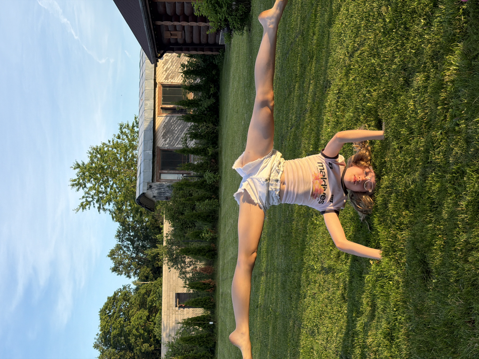 In Dreiliņi, Latvia, a young person is performing a handstand on a grassy lawn with their legs spread wide in a straddle split. They are wearing white denim shorts and a t-shirt with "Marines" printed on it in reverse. The background features a long, low building with a tin roof, a wooden log cabin, and a wall made of concrete blocks. Trees and bushes surround the area, and the sky above is a clear blue with some wispy clouds. The lighting suggests it is late afternoon or early evening, with the sun casting long shadows.