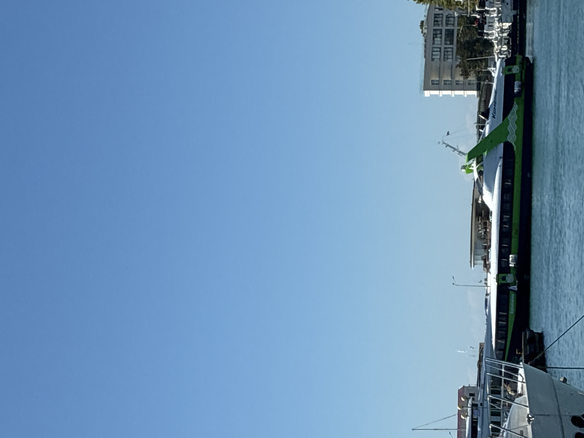 A bright, clear day with a vast blue sky dominates the upper portion of the image. In the lower half, a harbor scene unfolds with several boats. A prominent green and white hydrofoil, labeled "Комета" (Kometa), is docked. To its left, another white boat with a dark hull is partially visible, and further to the left, the bow of a white vessel is in the foreground. In the background, a modern, multi-story building with large windows stands behind the boats. The water appears calm. The scene is consistent with a maritime setting in Sochi, Russia, likely a port or marina. There are no visible individuals engaged in specific actions or interactions within the frame.