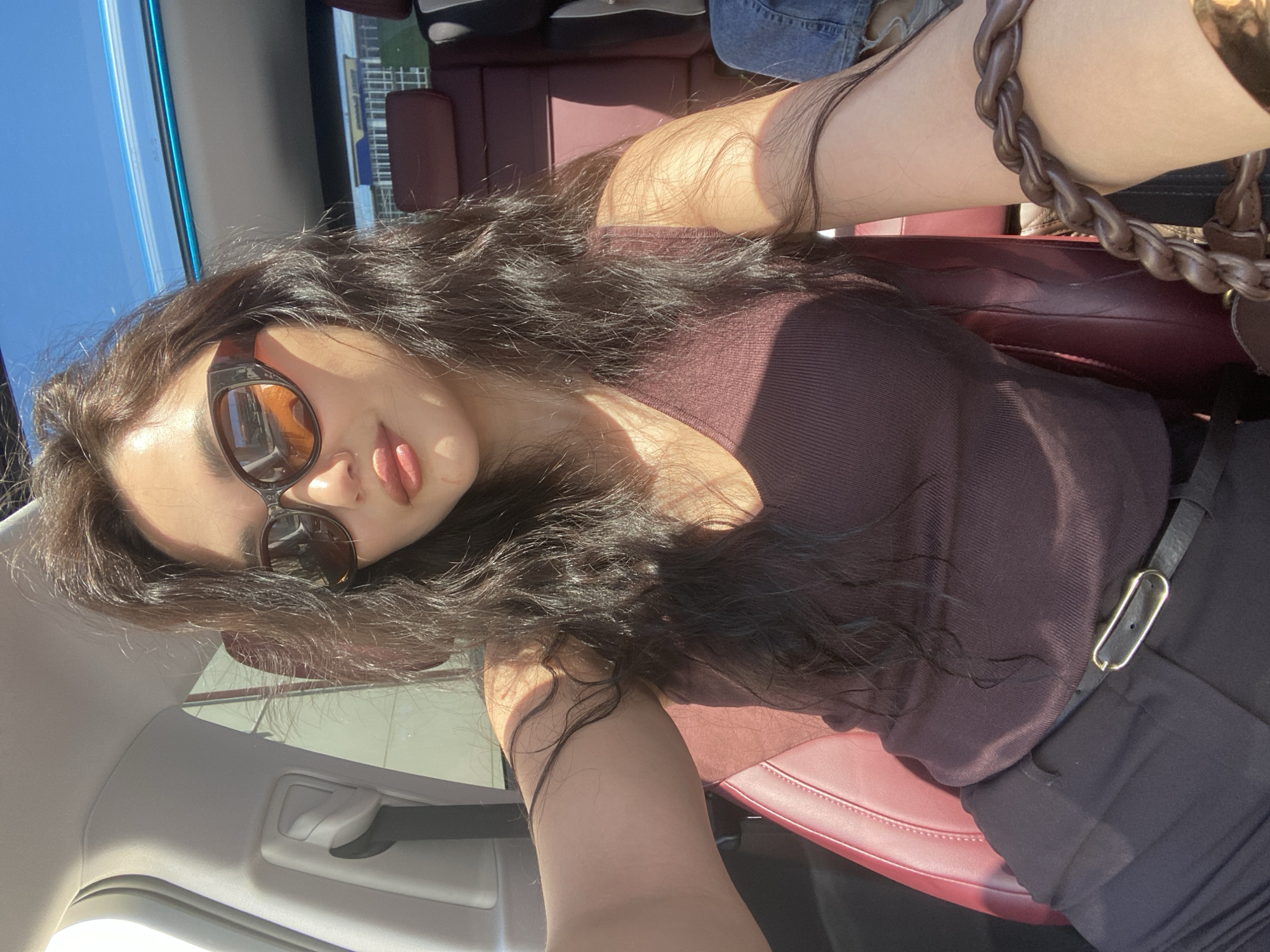 A casual and sunny self-portrait of a young woman taken inside a car in Sochi, Russia. The image is rotated, showing the woman from an overhead perspective, looking directly at the camera with a playful expression, her tongue slightly extended.
She has long, dark, wavy hair and is wearing stylish cat-eye sunglasses with dark frames and amber-tinted lenses. Her lips are adorned with pink lipstick. Her attire includes a dark brown or burgundy ribbed top and dark trousers, cinched with a black belt featuring a sleek, rectangular gold-toned buckle. On her right wrist, a thick, braided brown leather-like bracelet or bag strap is visible.
The car's interior features rich red/burgundy leather seats. Parts of the light-colored car ceiling and a grey door handle are also visible. The background, seen through the car window, reveals a clear blue sky and distant modern buildings, suggesting an urban setting on a bright, sunny day. Strong natural sunlight illuminates the scene, casting highlights on her hair and arms. The atmosphere is relaxed and stylish. There is no visible text in the image.
