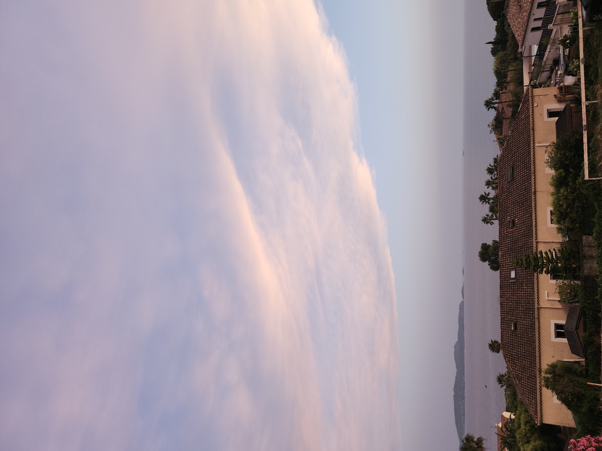 Captured at a sideways angle, the image reveals a picturesque coastal scene in Carqueiranne, France. Dominating the left is a pastel-colored sky, transitioning from light blue to soft pink with wispy cloud formations. To the right, a row of buildings, characterized by terra-cotta tile roofs and cream-colored walls, suggests residential architecture, with glimpses of windows and lush greenery. The foreground features palm trees and verdant foliage, hinting at a Mediterranean landscape. A body of water, likely the Mediterranean Sea, lies between the buildings and the sky, with distant landmasses barely visible through the hazy atmosphere.