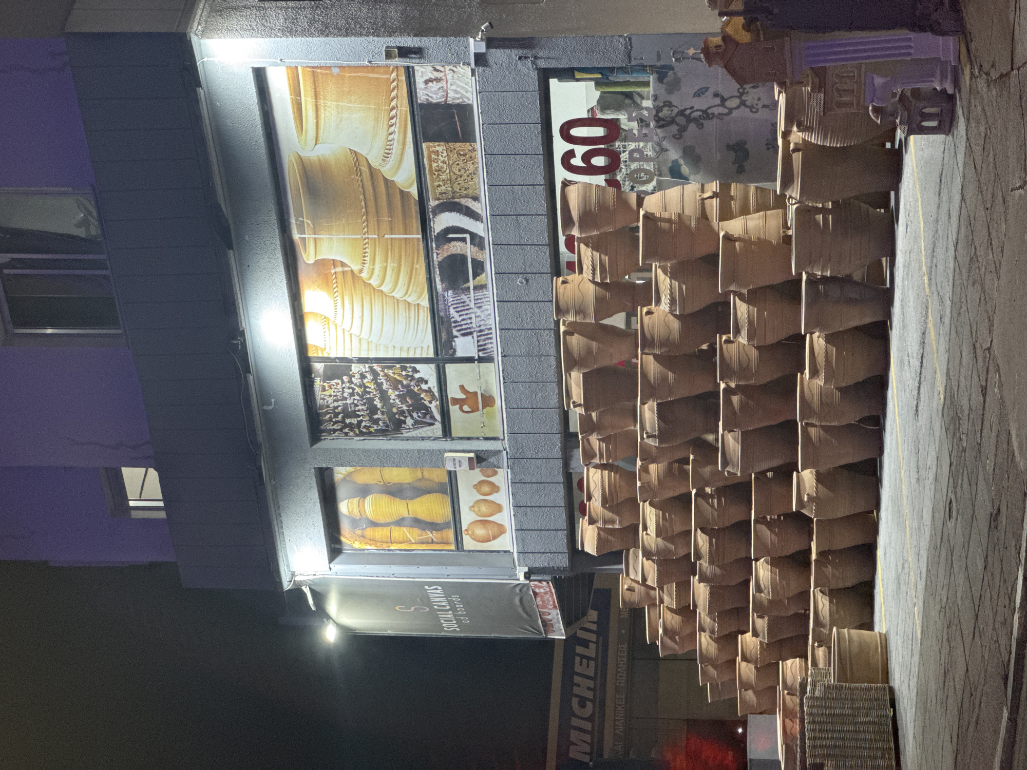 The image depicts a nighttime scene outside a shop in Limassol, Cyprus, specializing in pottery. The shop's facade features large windows displaying images of terracotta pots and other decorative items, including a sign for "SOCIAL CANVAS ad boards". A prominent "MICHELIN" sign is visible on the left, suggesting proximity to a tire shop or a business associated with the brand.
The foreground is dominated by a large, tiered display of numerous terracotta pots of various sizes and designs, stacked in a pyramid formation. Some smaller pots and a woven basket are also present. The ground is paved with cobblestones and marked with yellow lines.
Streetlights and internal shop lights illuminate the scene, creating stark contrasts and shadows. The building itself appears to be a multi-story structure with a balcony or upper floor visible. The overall impression is that of a commercial area at night, with the pottery display being the central focus. There are no people visible in the image.