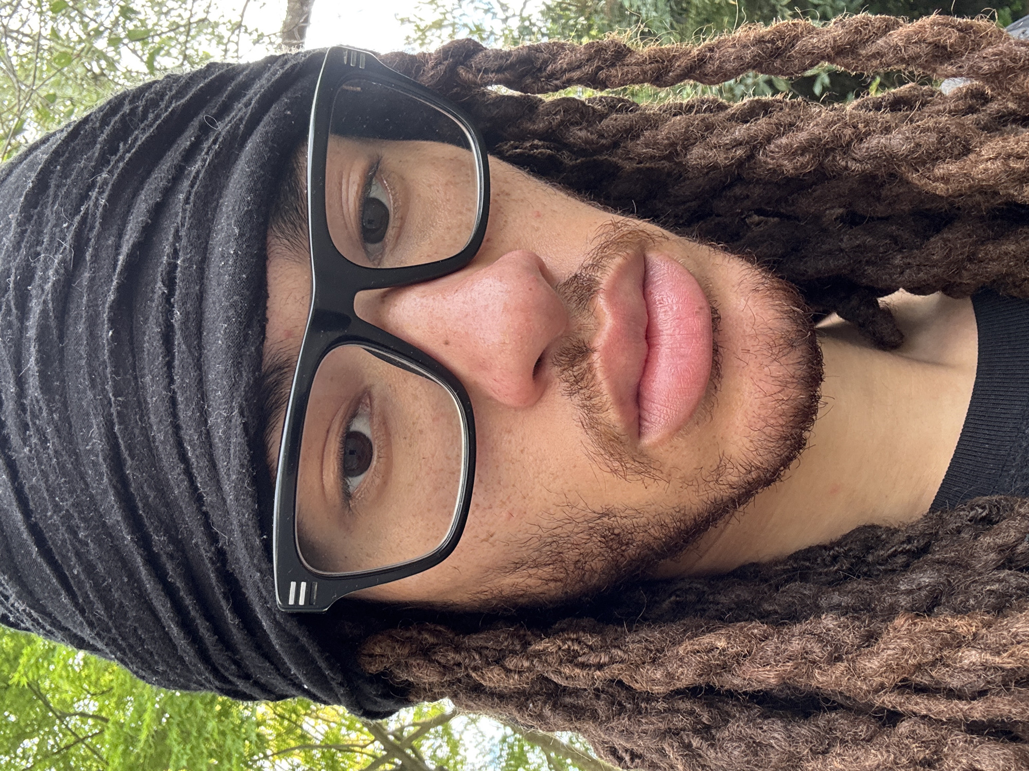 A close-up, tilted portrait of a person with brown dreadlocks and a black head wrap. They are wearing black-framed glasses and have light skin with a goatee. The background shows hints of trees and sky. There is no information to indicate the photo was taken in Les Ulis, France.