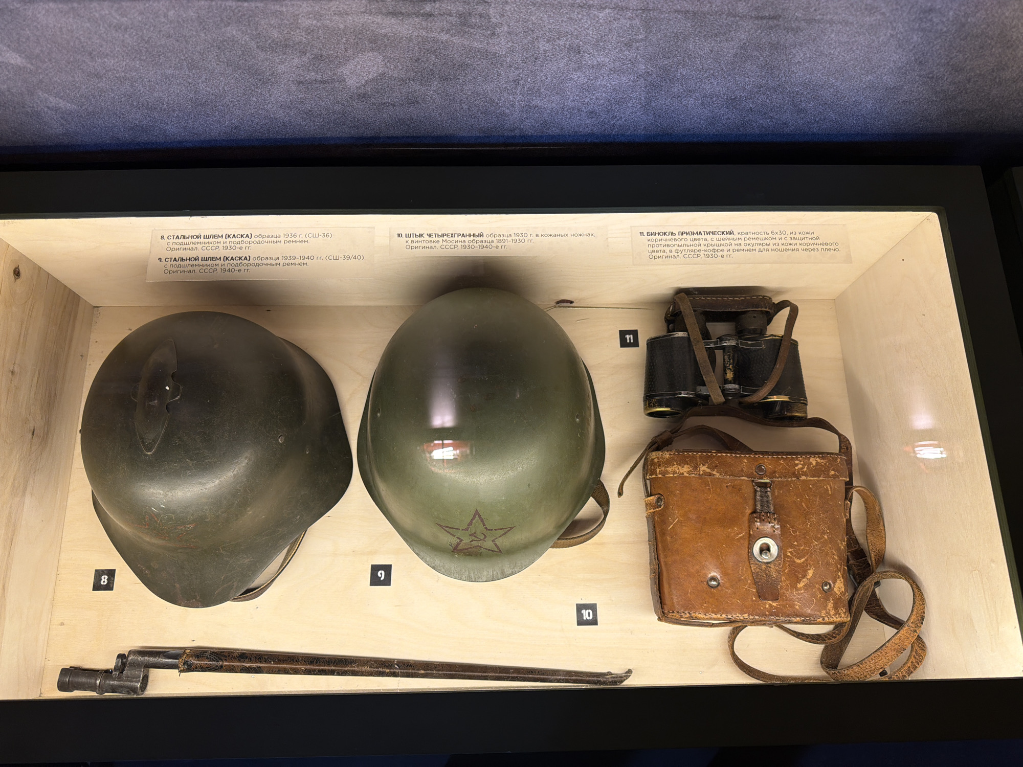 The image shows a display case containing military artifacts, likely in a museum in Krasnoyarsk, Russia. The artifacts are arranged neatly on a light-colored wooden surface. The artifacts include:
1. A dark green steel helmet (item 8), identified by a label as a model from 1936 (SSH-36), complete with liner and chinstrap.
2. A lighter green steel helmet (item 9), labeled as a model from 1939-1940 (SSH-39/40), also with liner and chinstrap. It has a painted red star on the front.
3. A bayonet (item 10), described as a four-sided model from 1930, in leather scabbard. It is associated with the Mosin rifle model 1891-1930.
4. A pair of 6x30 prismatic binoculars (item 11), brown leather case with a neck strap, protective anti-dust cover for the eyepieces, and carrying strap.
Each artifact is labeled with a number and corresponding information about its type, origin, and period of use. The environment is indoors, with artificial lighting illuminating the display.