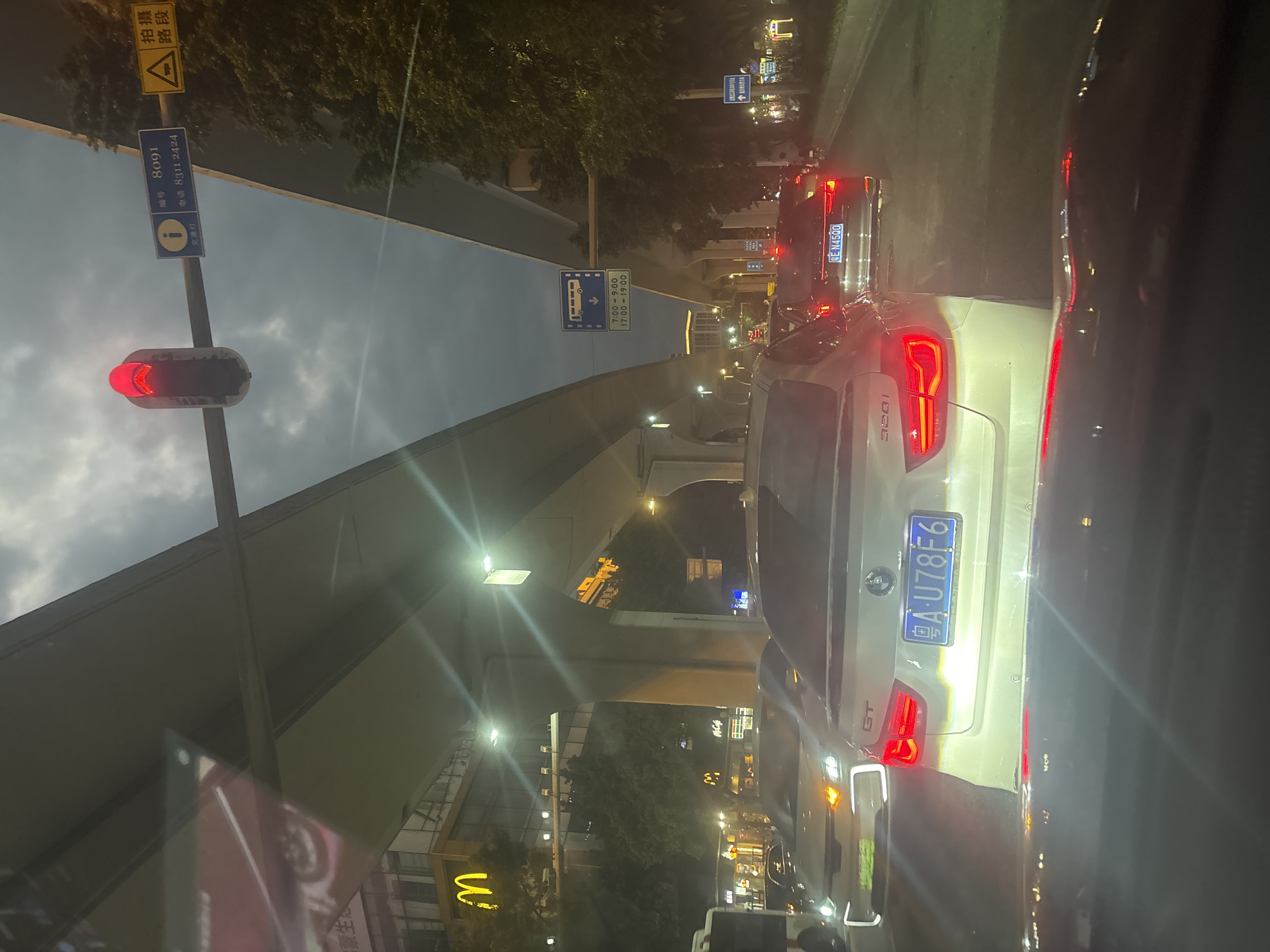 The image depicts a traffic scene at night in Baiyun District, China, likely near a McDonald's restaurant judging by the visible signage. The sky is dark and cloudy, with artificial lights illuminating the road and surrounding buildings. A traffic light is visible, displaying a red signal. Several cars are present on the road, stopped in traffic. The foreground shows the interior of a vehicle, with the dashboard and windshield visible. In the background, an elevated road structure casts shadows over the scene. A McDonald's sign is prominently displayed on a building to the left. Various other signs and lights are visible, indicating a commercial and urban environment. The overall atmosphere is that of a busy city street at dusk or night.