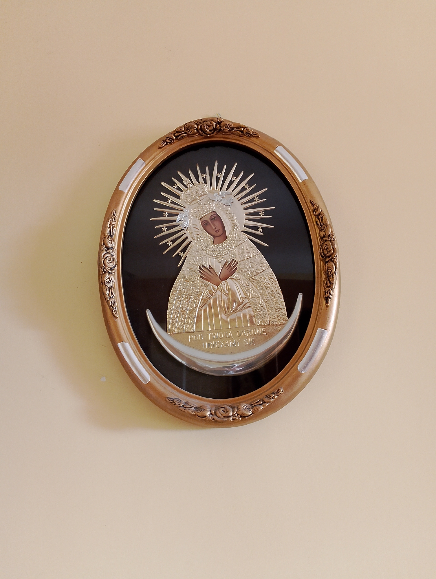 The image shows a framed icon of the Virgin Mary hanging on a light beige wall. The icon is oval-shaped with a gold-colored frame featuring floral motifs. The Virgin Mary is depicted with a halo and a crown, wearing a richly ornamented robe. She is standing on a crescent moon, and her hands are clasped in front of her chest. Below her image, there is text in Polish, which reads "POD TWOJĄ OBRONĘ UCIEKAMY SIĘ". This translates to "Under your protection, we flee". The icon is centrally located on the wall, with ample empty space around it. The lighting appears to be natural, coming from the right side of the frame, casting a slight shadow on the left. The scene suggests a religious or devotional setting, possibly a home or a church.
The image likely originates from Kolno, Poland, as indicated by the Polish text on the icon, which is a common devotional phrase associated with the Black Madonna of Częstochowa, a revered icon in Poland. While the specific location within Kolno cannot be determined from the image alone, such icons are typically found in churches, chapels, or private residences in Polish towns and cities.