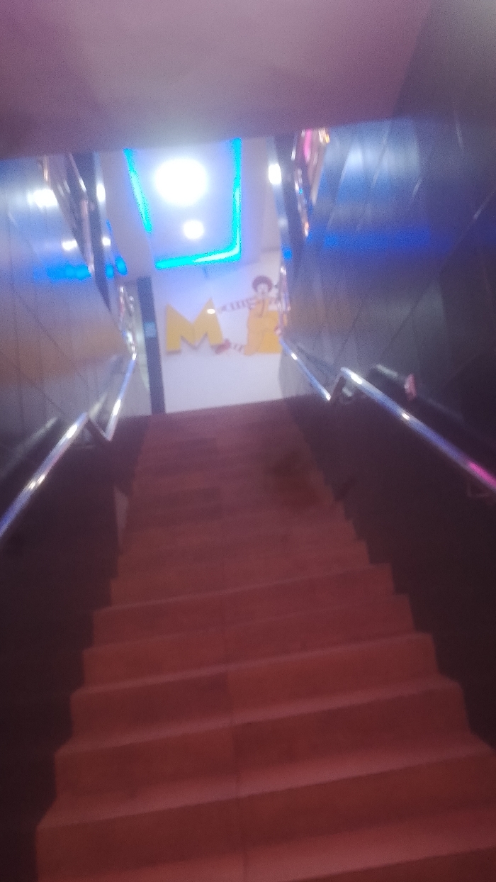 A blurred perspective of red stairs leading upwards. Metal handrails flank both sides. At the top of the stairs is a wall featuring a large yellow "M" and a cartoon figure. Blue and white lights accent the ceiling above the wall. The surrounding walls appear dark, adding to the low-light ambiance.