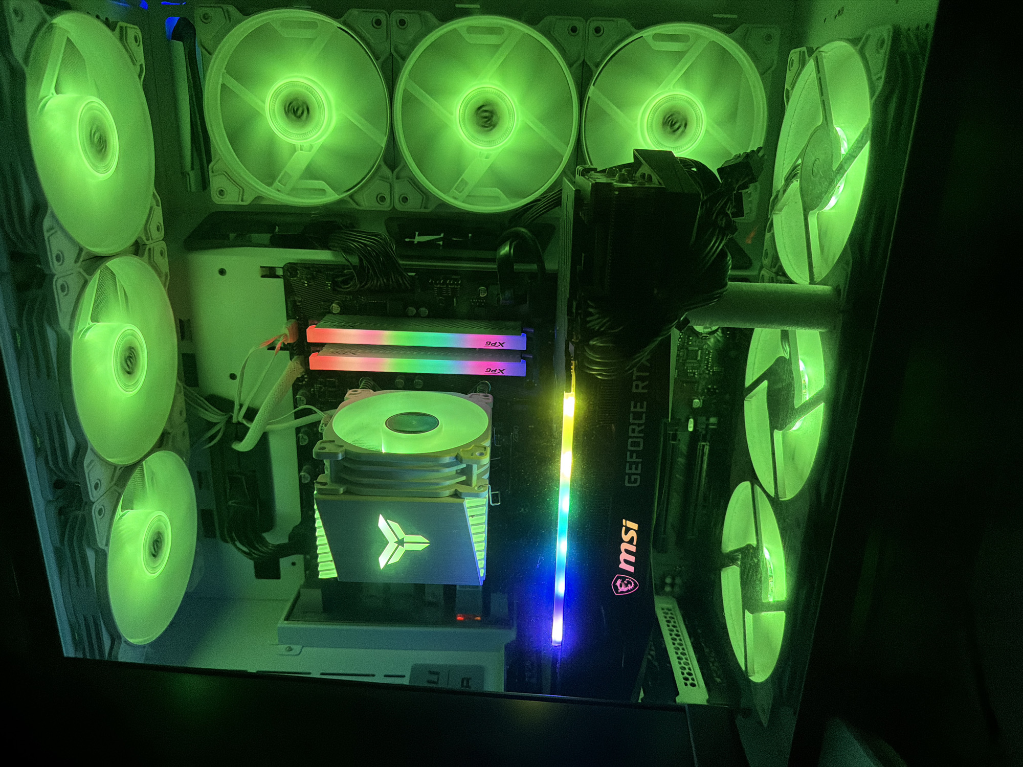 The image shows the interior of a computer tower with several components visible, all illuminated by bright green LED lights from multiple fans. The computer's components include a CPU cooler with a stylized logo, RAM sticks with RGB lighting, and a graphics card with "msi GEFORCE RTX" branding. The fans are mounted at the top, side, and bottom of the case, and their lighting creates a vibrant green glow throughout the interior. The overall scene is dark, with the exception of the bright green illumination from the computer's internal lighting. There are no people or external environmental elements visible in the image. The location is unknown.