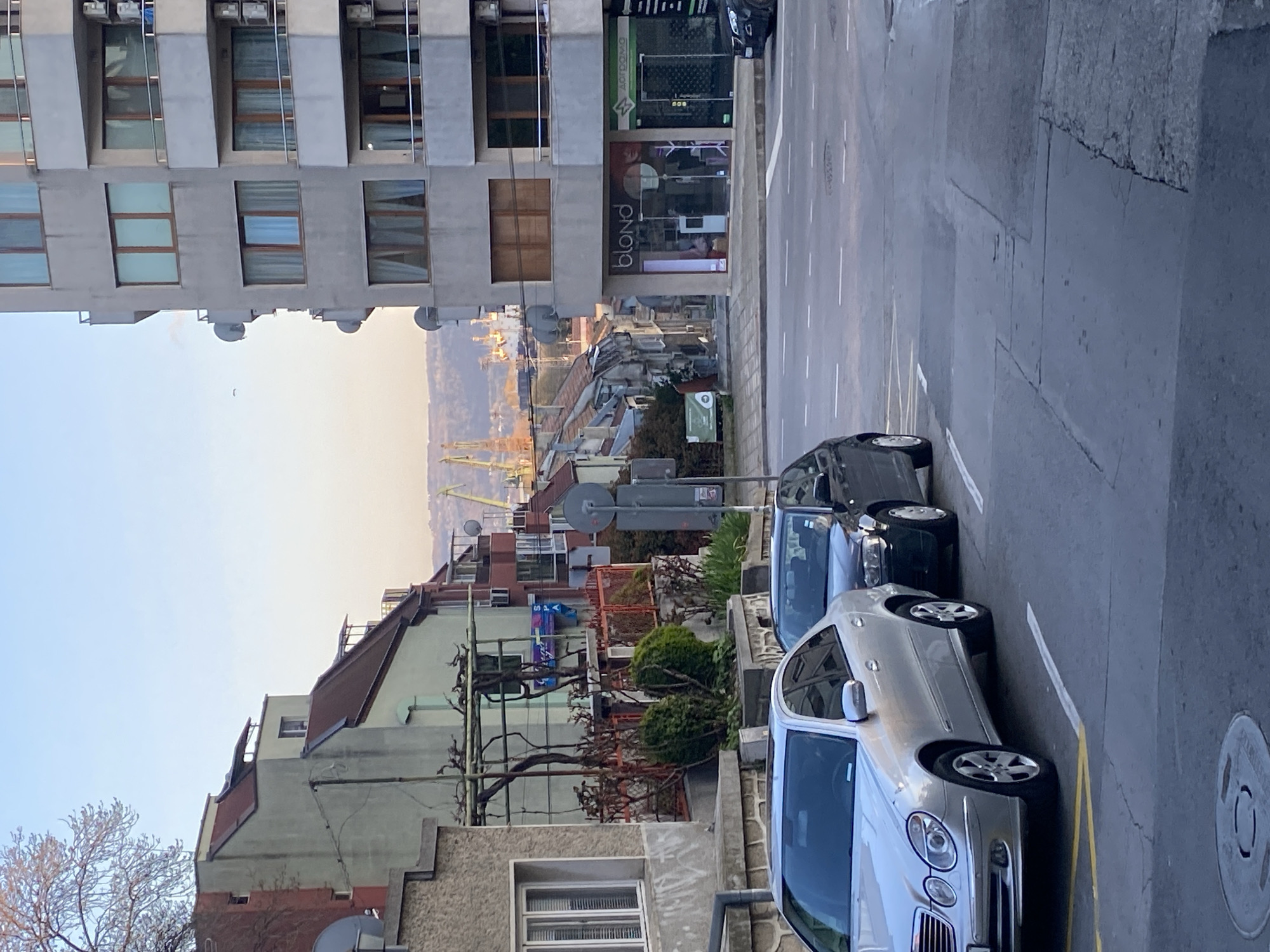 This image captures an urban street scene in Varna, Bulgaria, viewed from an elevated perspective looking down a street towards a distant harbor and hills.
In the foreground, a paved street with distinct white and yellow lane markings is visible. Several cars are parked along the right curb, including a silver Mercedes-Benz sedan and a dark-colored sedan behind it. A circular metal manhole cover is on the sidewalk in the lower right.
To the left, a multi-story modern building with a concrete facade and numerous rectangular windows, some partially obscured by curtains, stands prominently. Its ground floor houses a commercial establishment with a large glass storefront. Visible text on the storefront includes "blond" in white stylized letters, likely indicating a beauty salon or similar business. Above "blond," Cyrillic text is present, including "ДОРОГАТА" (The Road).
Beyond this main building, a dense collection of other urban structures, primarily residential, extends into the midground. These buildings feature varying architectural styles, with visible details like satellite dishes, balconies, and possibly clotheslines. Bare tree branches are seen in the lower left, and some green shrubs dot the midground landscape. A street sign pole with a circular sign is visible near the parked cars.
In the far background, under a clear, light blue sky, a body of water (likely the Black Sea, given the Varna context) stretches out, bordering a port or industrial area identifiable by several tall yellow cranes. Gentle hills rise beyond the port in the distance.
The time of day appears to be early morning or late afternoon, characterized by soft, natural lighting that casts a slight warmth on the distant landscape. The weather is clear and calm. The scene depicts a quiet moment in an active urban environment, with no specific event or activity occurring.