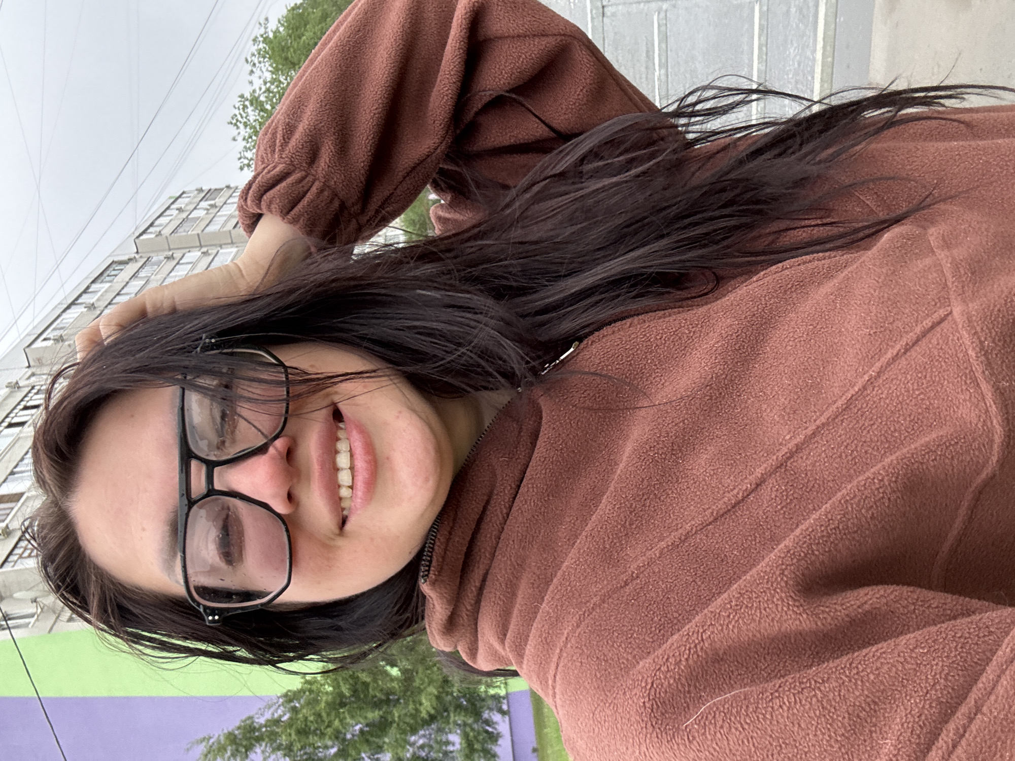 A woman with dark hair and glasses smiles at the camera while standing outside. She is wearing a brown, long-sleeved fleece jacket. Her left hand is positioned on her head. Behind her, there is a multi-story building and some trees. The buildings in the city, Unknown, Unknown, are visible in the background. The weather appears overcast.