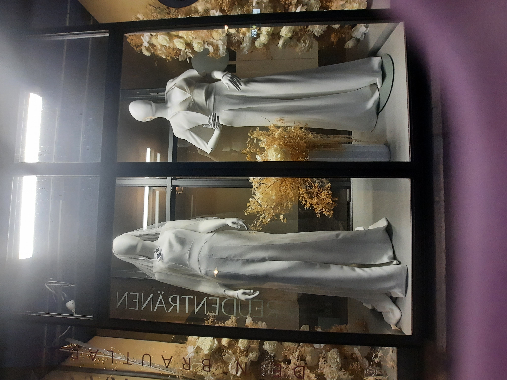 The image shows a store window display featuring two mannequins dressed in white bridal attire. The mannequin on the left wears a long, flowing wedding dress with a veil. The mannequin on the right wears a white jumpsuit with a matching jacket. The display is decorated with floral arrangements, including white roses and dried grasses. A sign on the left side of the window, partially obscured, reads "DEBRA RAUTILA" in gold lettering. The store appears to be located in Rosenheim, Germany, as indicated by text on the window that is partially visible and reversed, reading "BRAUTMODE ROSENHEIM" which translates to "bridal fashion Rosenheim". The lighting in the window suggests it is nighttime or dusk.