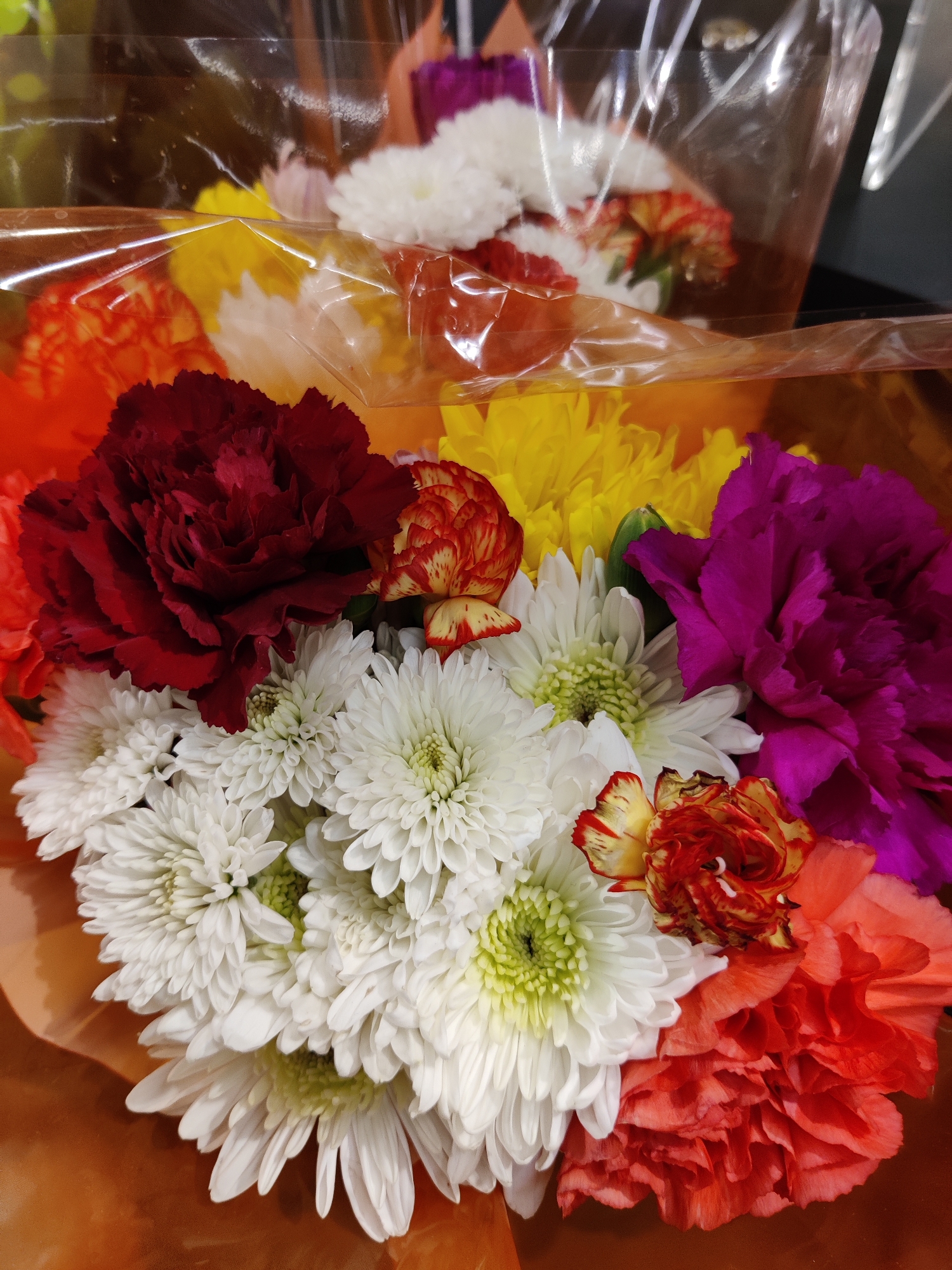 A close-up, high-angle shot shows a bouquet of vibrant flowers, predominantly white chrysanthemums, arranged with a deep red carnation, a bright pink carnation, yellow mums, orange carnations, and a few small, red and yellow flecked carnations. The flowers are enclosed in clear plastic packaging, with a sheen suggesting a retail display. The background is blurred, but hints at a dark surface. The bouquet appears to be a retail display item. The image does not contain any recognizable landmarks or context that would place it in Mechanicsburg, United States of America.