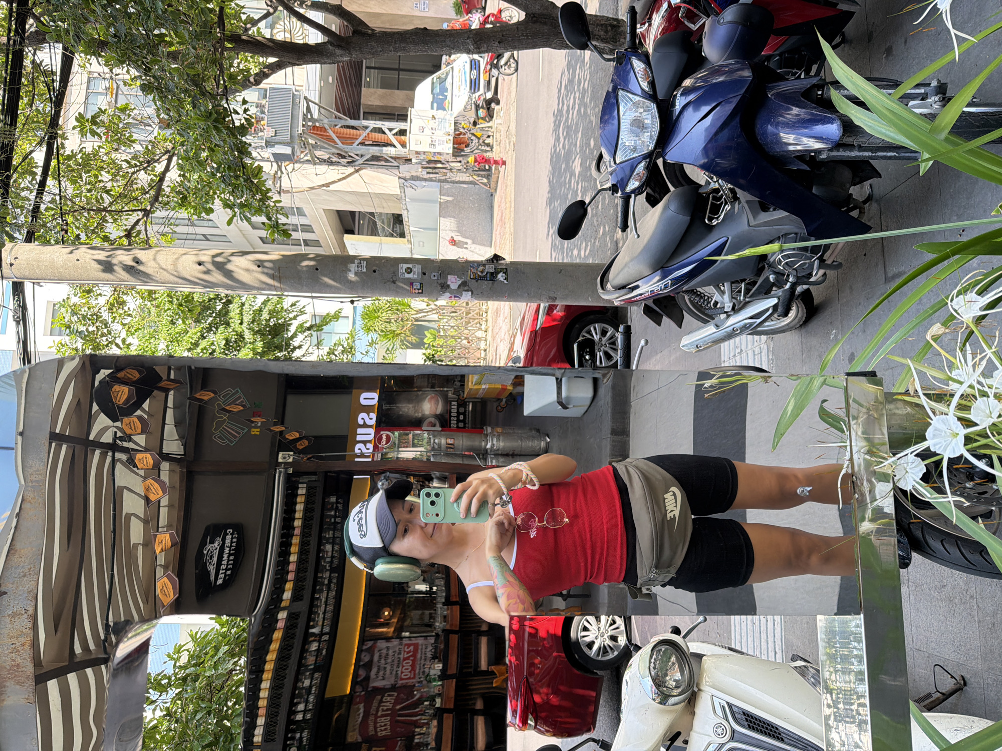 A woman stands on a sidewalk in front of a mirrored storefront in Nha Trang, Vietnam, taking a selfie. She is wearing a red tank top, black shorts, a green fanny pack, a baseball cap, and large green headphones. She holds a green smartphone in her right hand. In the mirror, her reflection shows her and the street scene behind her. The street is lined with trees and buildings. Several motorcycles are parked along the sidewalk. A red car is visible in the distance. The storefront appears to be a bar or restaurant with a display of bottled drinks.