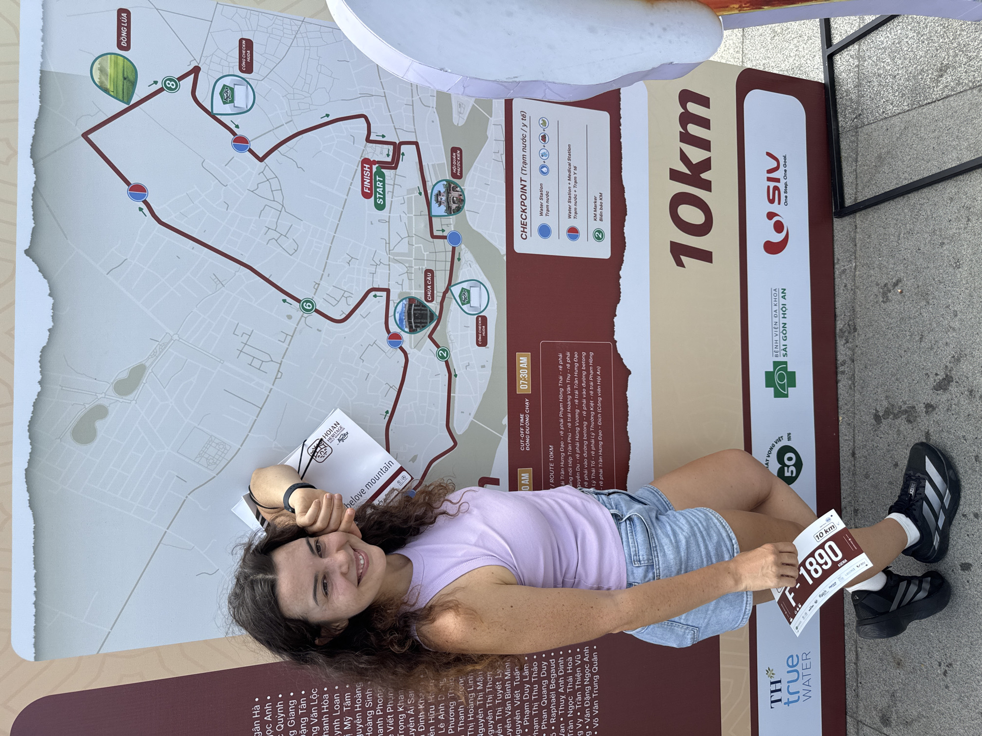 A young woman with curly brown hair and a light purple tank top and denim shorts stands in front of a large map depicting a 10km race route in Hoi An, Vietnam. She is smiling and holding a race bib with the number F-1890. The map shows various checkpoints and landmarks, including "CHUA CAU" (Japanese Covered Bridge) and "HOI QUAN PHUC KIEN" (Fujian Assembly Hall). The race route is marked in red with numbered circles indicating checkpoints. The overall scene suggests an outdoor event or information booth related to a running race in the historic city.
