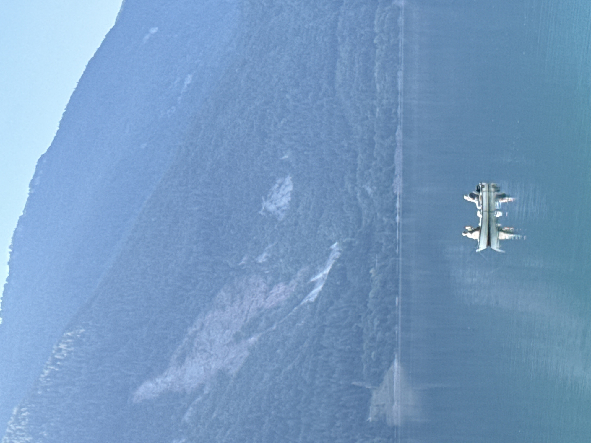 In Molveno, Italy, a wide shot captures a serene, calm, bright blue lake reflecting the sky and a verdant mountain slope under a hazy, pale blue sky. In the middle of the lake, a small white rowboat with two occupants is visible. The boat is oriented diagonally, with the two figures positioned toward the back. One figure is seated, and the other is standing. The boat is creating a faint reflection on the water's surface. The mountain, covered in dense green trees, rises steeply from the far shore of the lake, with a lighter-colored streak running down its side. The overall scene is peaceful and natural.