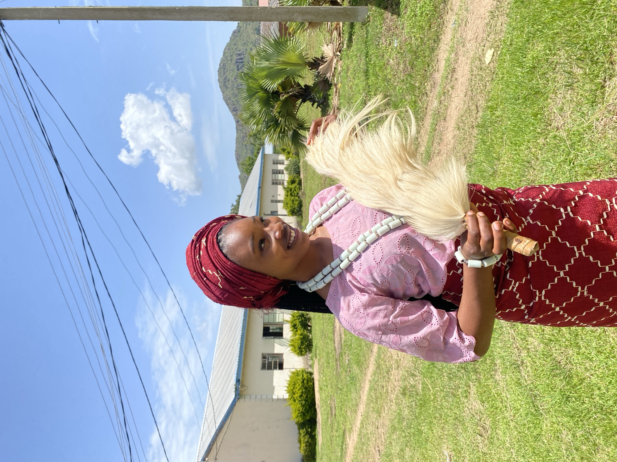 An African woman stands outside, wearing a traditional red headwrap and a pink patterned blouse, holding a tan horsetail whisk in her right hand. She is adorned with multiple strands of light-colored beads around her neck and a white bracelet on her right wrist. Her skirt features a dark red fabric with a repeating geometric white pattern. In the background, a light-colored building with a metal roof and several windows sits on a grassy expanse, with green bushes flanking it. Beyond the building, a lush green hill rises against a bright blue sky dotted with white clouds. The scene is likely located in Vom, Nigeria, given the architectural style, vegetation, and overall landscape often found in the region.