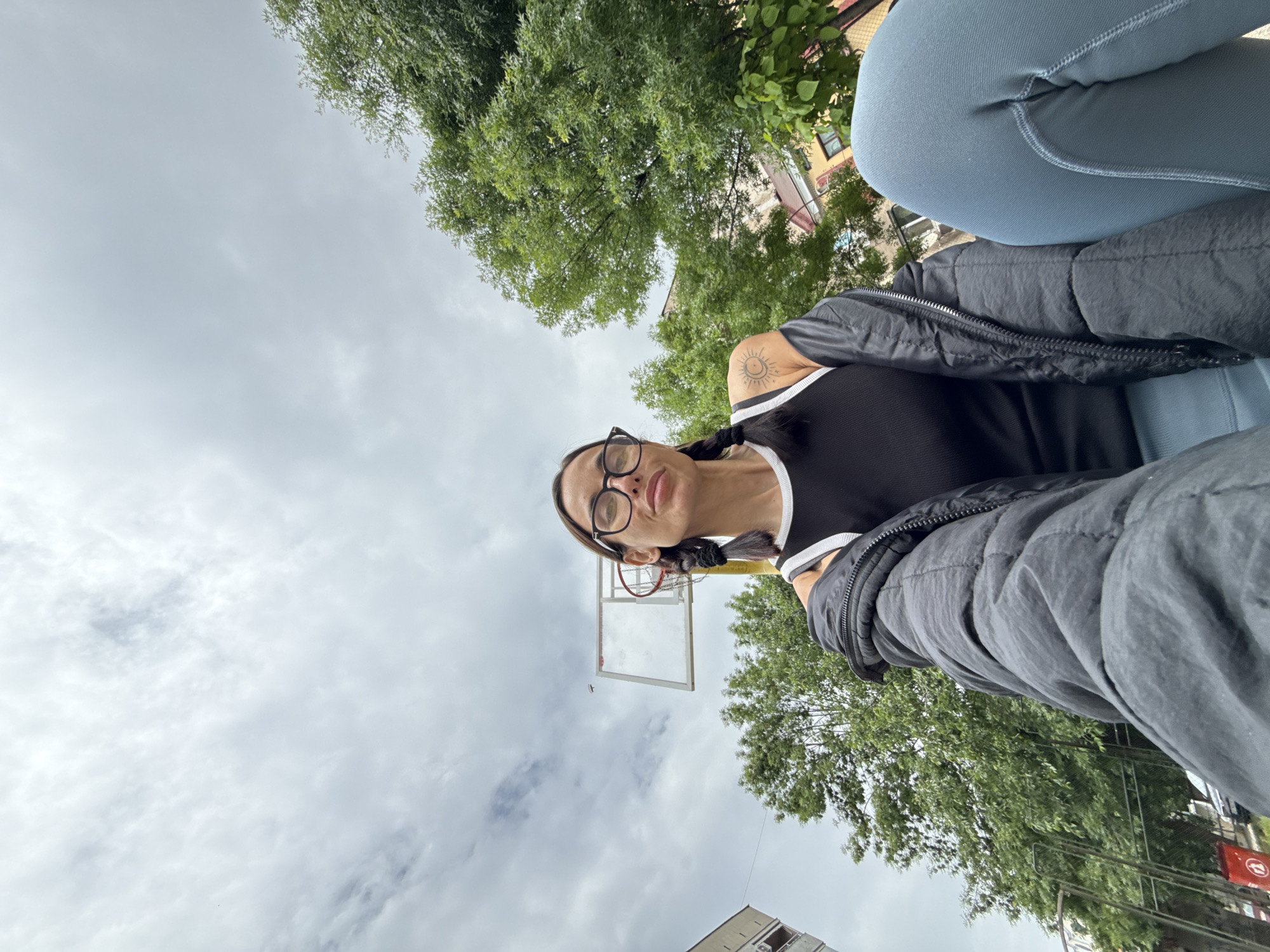 A low-angle, selfie-style photo shows a woman with dark hair in braids, wearing glasses, a black and white tank top, a dark jacket, and light blue leggings. She's positioned against a backdrop of cloudy sky and green trees, with a basketball hoop visible behind her head. The setting appears to be an outdoor court or park. A building is visible at the bottom left. The location is Pleven, Bulgaria.