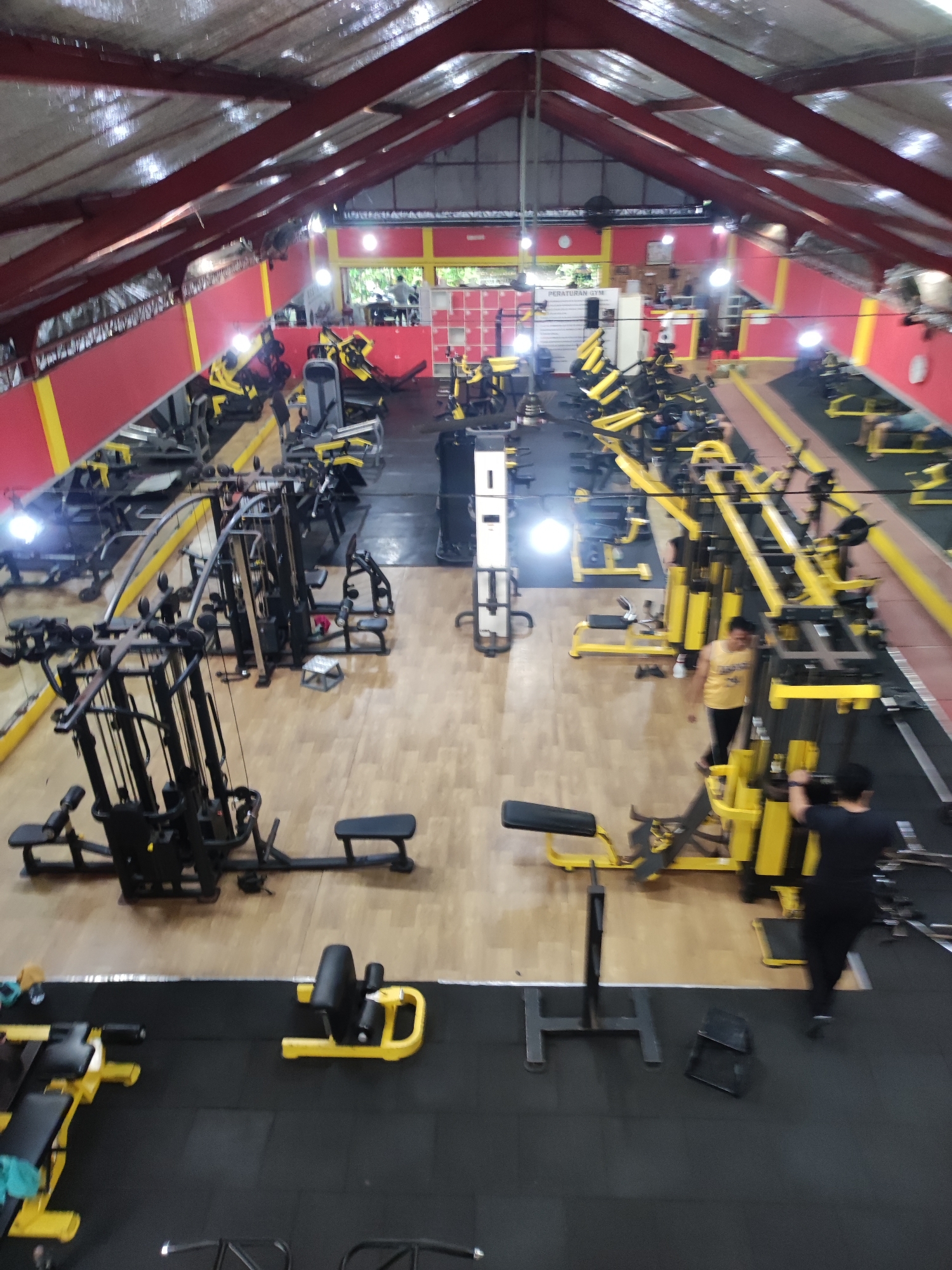 The image depicts the interior of a gym located in Kedewatan, Indonesia. The gym is well-equipped with various exercise machines, most of which are colored black and yellow. The flooring consists of both light-colored wood and black rubber matting. There are a few individuals present, including people exercising on the machines and moving throughout the gym. The walls are painted red with yellow trim, and the roof has an industrial appearance with exposed metal beams. A set of gym rules is posted in the upper part of the image.