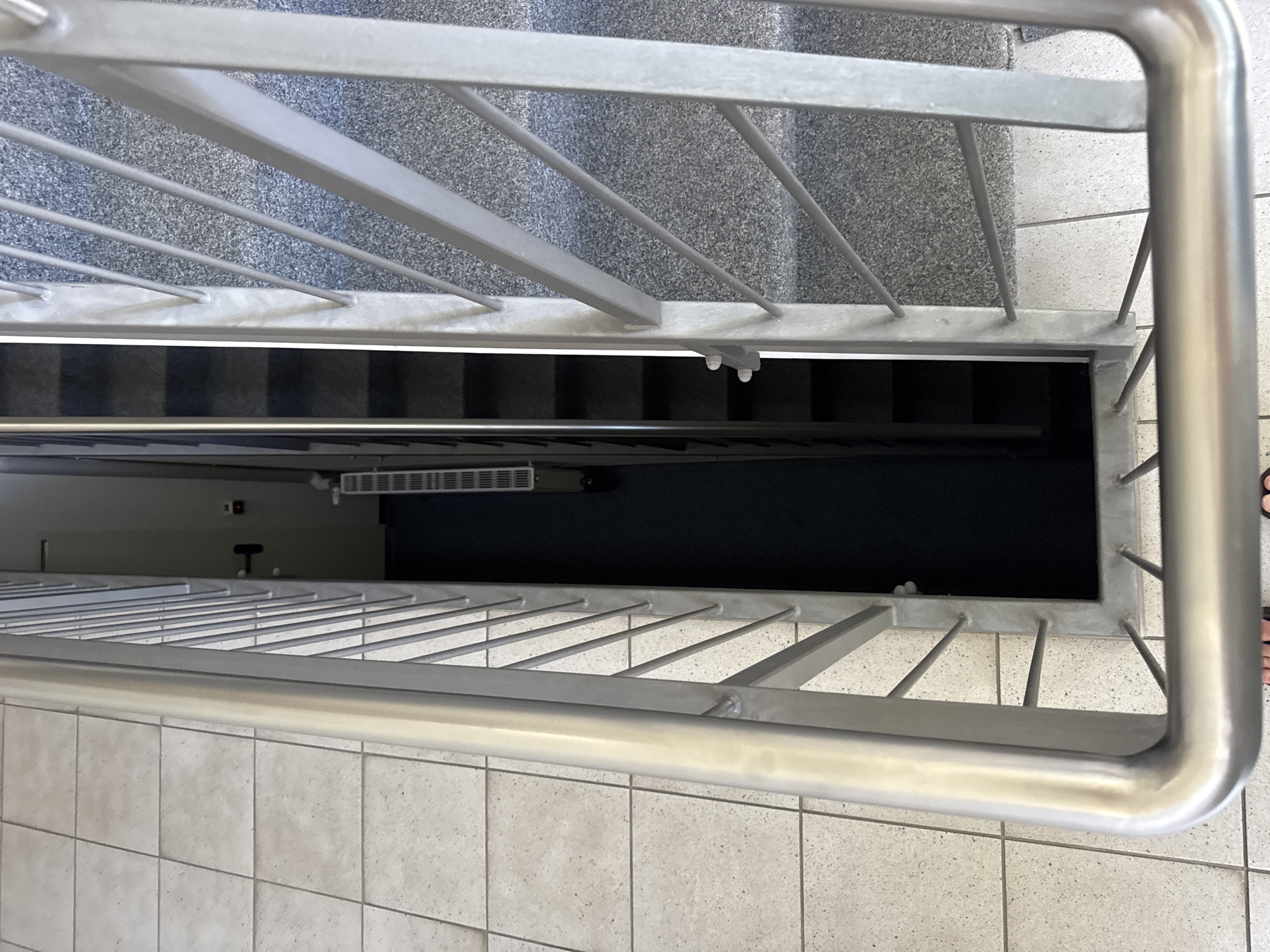 This is a top-down shot of a stairwell in Höhenkirchen-Siegertsbrunn, Germany, likely in a public building or apartment complex. The foreground features a polished metal handrail curving to the right, with visible welds. To the left, a tiled floor with light gray square tiles and darker grout lines extends outwards. The stairwell itself is visible beyond the railing, featuring dark gray carpeted steps and a speckled gray material on the landings. On the left side of the stairwell, a white radiator is mounted on the wall. Further down the stairwell, a dark blue carpet covers the floor. A black door is visible in the background. The scene is brightly lit, suggesting an indoor environment during daylight hours.