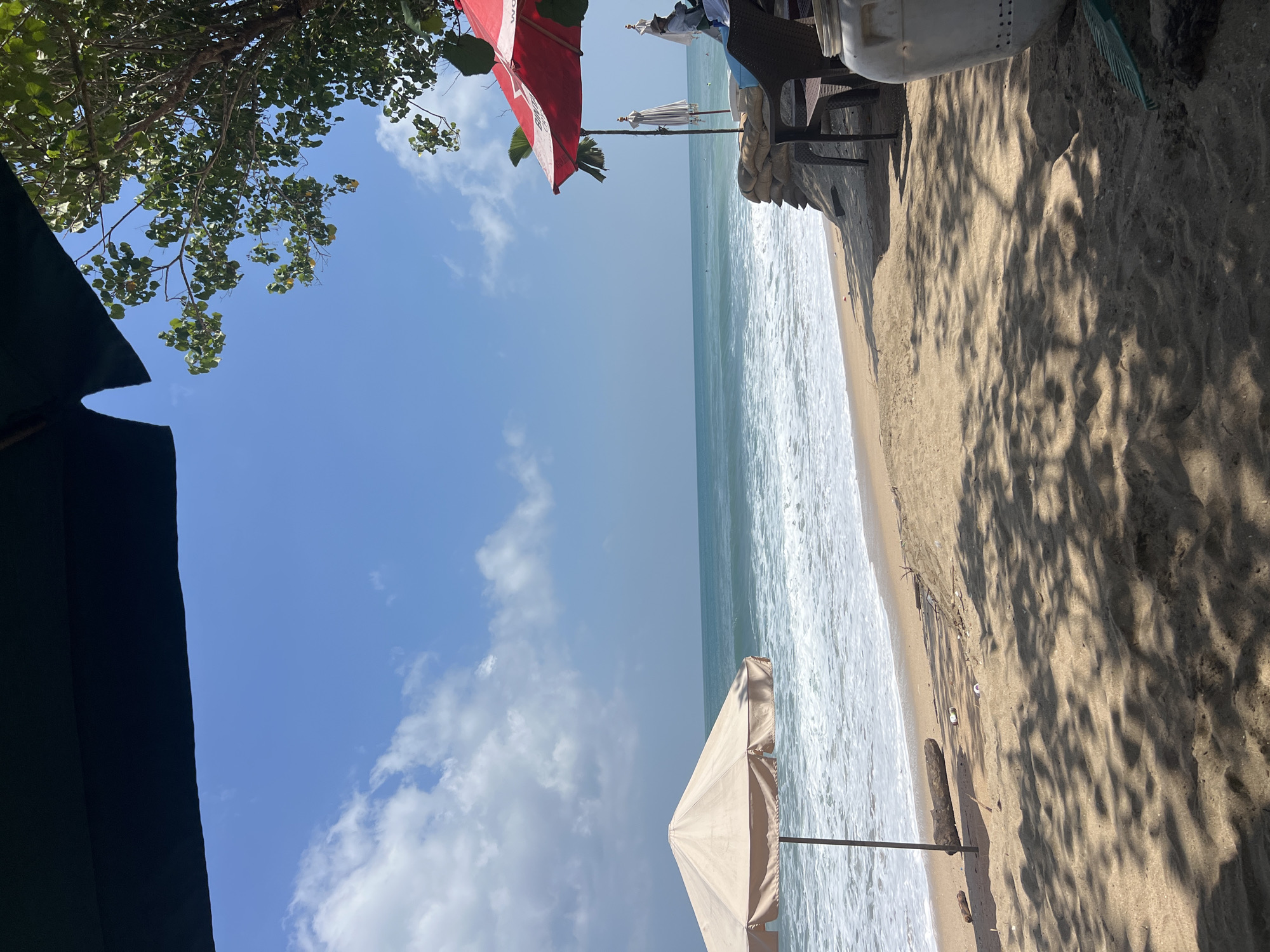 The image depicts a sunny beach scene in Kuta, Indonesia. The foreground is covered in sand with dappled shade from overhead foliage. A light-colored beach umbrella stands on the left, angled towards the ocean. Beyond the sand is the sea, with gentle waves breaking on the shore. In the middle ground, a row of sandbags forms a barrier, and a table and chairs are set up beneath red and white umbrellas. A person in a straw hat is seated in one of the chairs. A large white plastic container with a green rake rests on the sand to the right of the frame. The sky is blue with scattered white clouds. The overall impression is of a relaxed beachside establishment.