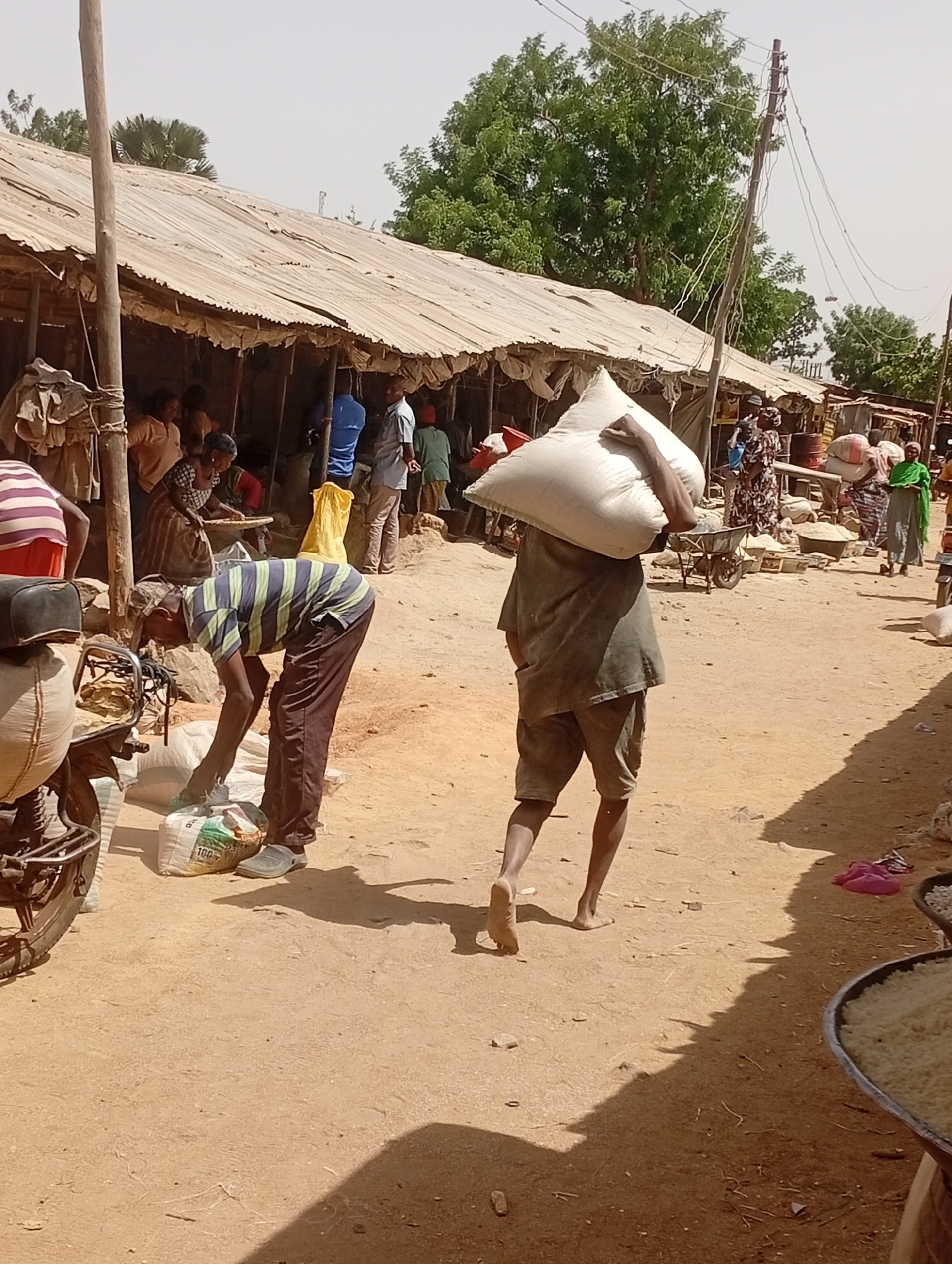 The image depicts a bustling market scene in Barnawa, Nigeria. A man in a striped shirt is bent over a bag on the ground near a parked motorcycle. Another man is walking barefoot with a large white sack over his shoulder. Other individuals are visible in the background near stalls under a corrugated metal roof, some using wheelbarrows. The ground is sandy and there are objects scattered around, including bowls or trays filled with grains. Overhead power lines are visible.