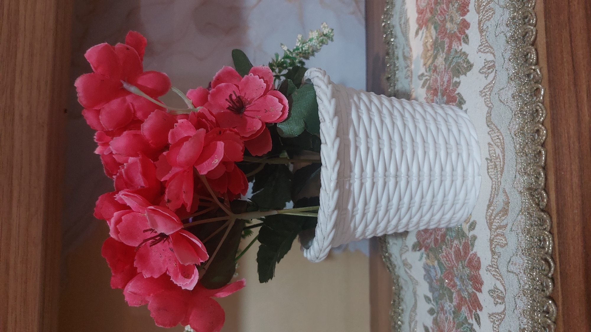 An artificial bouquet of bright pink flowers is displayed in a white, woven pot. The pot sits on a decorative table runner with a floral pattern and a gold, scalloped edge. The background is softly blurred, showing a marbled pattern and a wooden surface above. The image appears to be taken indoors, possibly in a home setting in the city of Medan, Indonesia. There are no people visible in the image.