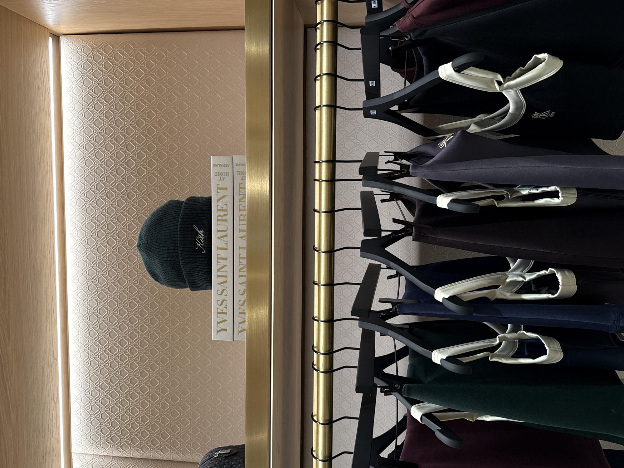 This image depicts an interior retail display at the Kith London flagship store, located at 18-20 Russell Street, Covent Garden, London WC2B 5EA. The scene is bisected horizontally by a brass-toned shelf and a brass clothing rail, set against a light beige wall panel featuring a subtle, repeating geometric heart-link pattern.
On the upper shelf, constructed of light wood with a brass-colored front trim, a dark green knitted beanie hat with "Kith" embroidered in white is prominently displayed. Behind the beanie, a stack of two white-bound books, identified by their spines as "YVES SAINT LAURENT" and "ASSOULINE AT HOME," are visible. A portion of a dark, textured object, possibly a bag, is visible on the far left. A linear LED light strip illuminates the patterned wall directly above this shelf.
Below, a brass-colored rail supports multiple black wooden clothing hangers, many bearing "KITH" branding near their hooks. These hangers display various items of casual or athletic apparel in a palette of dark colors including burgundy, dark green, navy blue, dark grey, and black. Several garments, appearing to be tops or tank tops, feature contrasting cream or white trim and visible "KITH" logos, such as a white logo on a black garment. No individuals are present in the frame.
