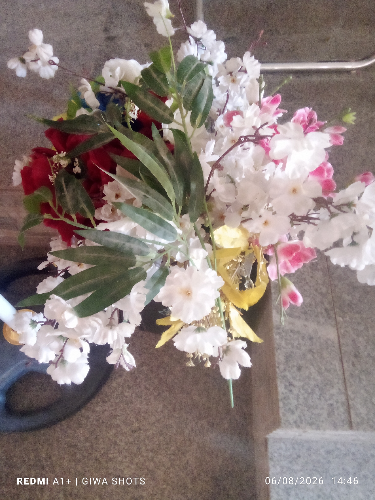 A top-down medium shot captures a bouquet of artificial flowers, predominantly white and pink with some red accents, arranged in a dark vase. The flowers, with delicate petals and green foliage, are densely packed. A bright yellow, ribbon-like accent is draped among the blossoms. The background consists of a textured, gray surface, possibly stone or concrete, with a hint of a light-colored ledge or step in the lower right corner. The image is overexposed in some areas, particularly around the lighter flowers, and shows a date and time stamp in the lower right corner: "06/08/2026 14:46", and a text overlay in the lower left corner: "REDMI A1+ | GIWA SHOTS". The lighting appears to be overhead and diffuse.