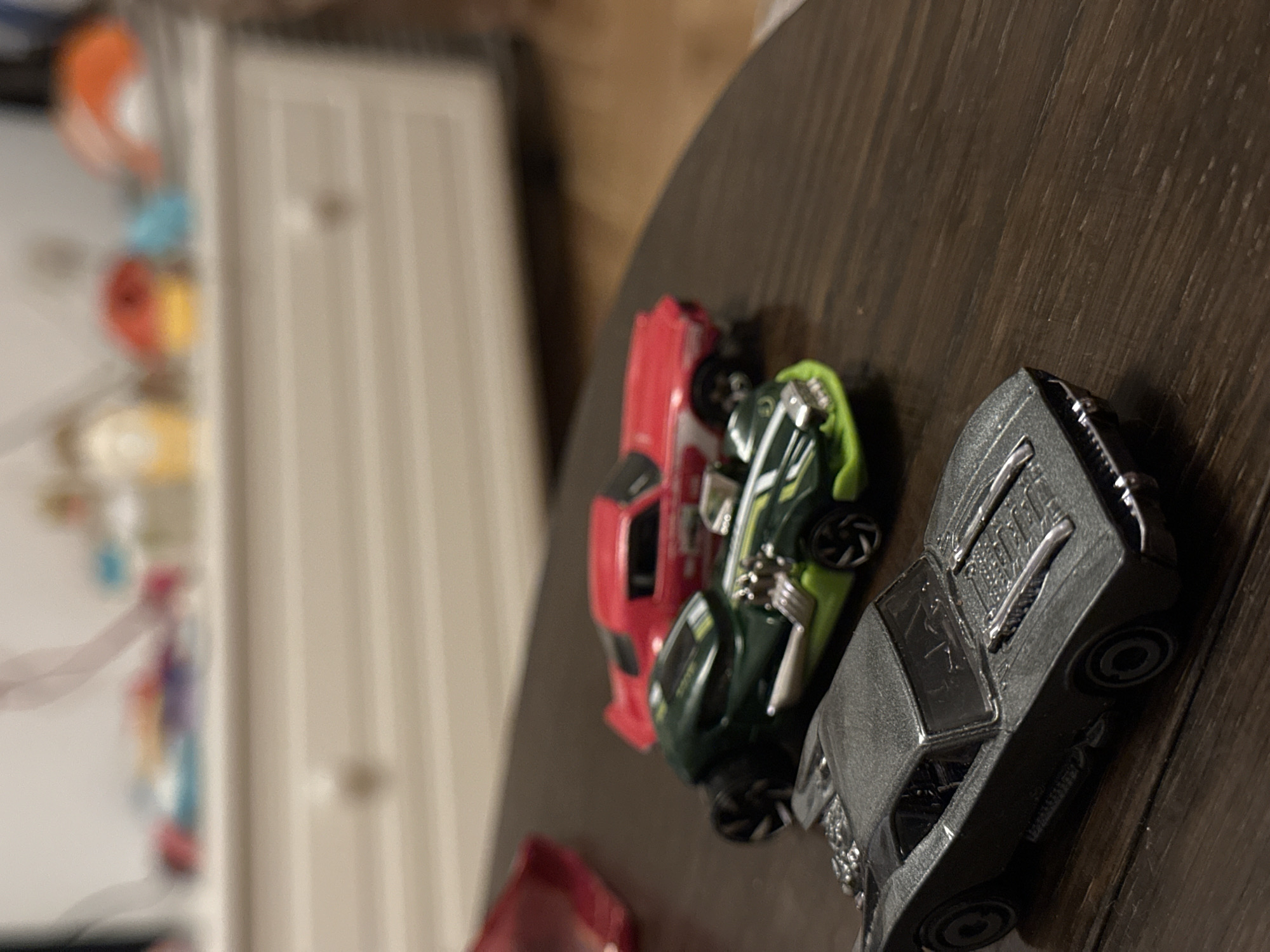 A close-up, eye-level shot shows three toy cars on a dark wooden surface. The cars are arranged in a line from closest to farthest. The closest car is a metallic gray Batmobile, with visible engine details on the hood and a prominent spoiler at the rear. Behind it is a dark green and lime green race car with chrome accents. The farthest car is red with white racing stripes. The background is blurred, showing a white dresser with drawers and some colorful objects on top. The scene is indoors, with no discernible view of the city of Sofia, Bulgaria.