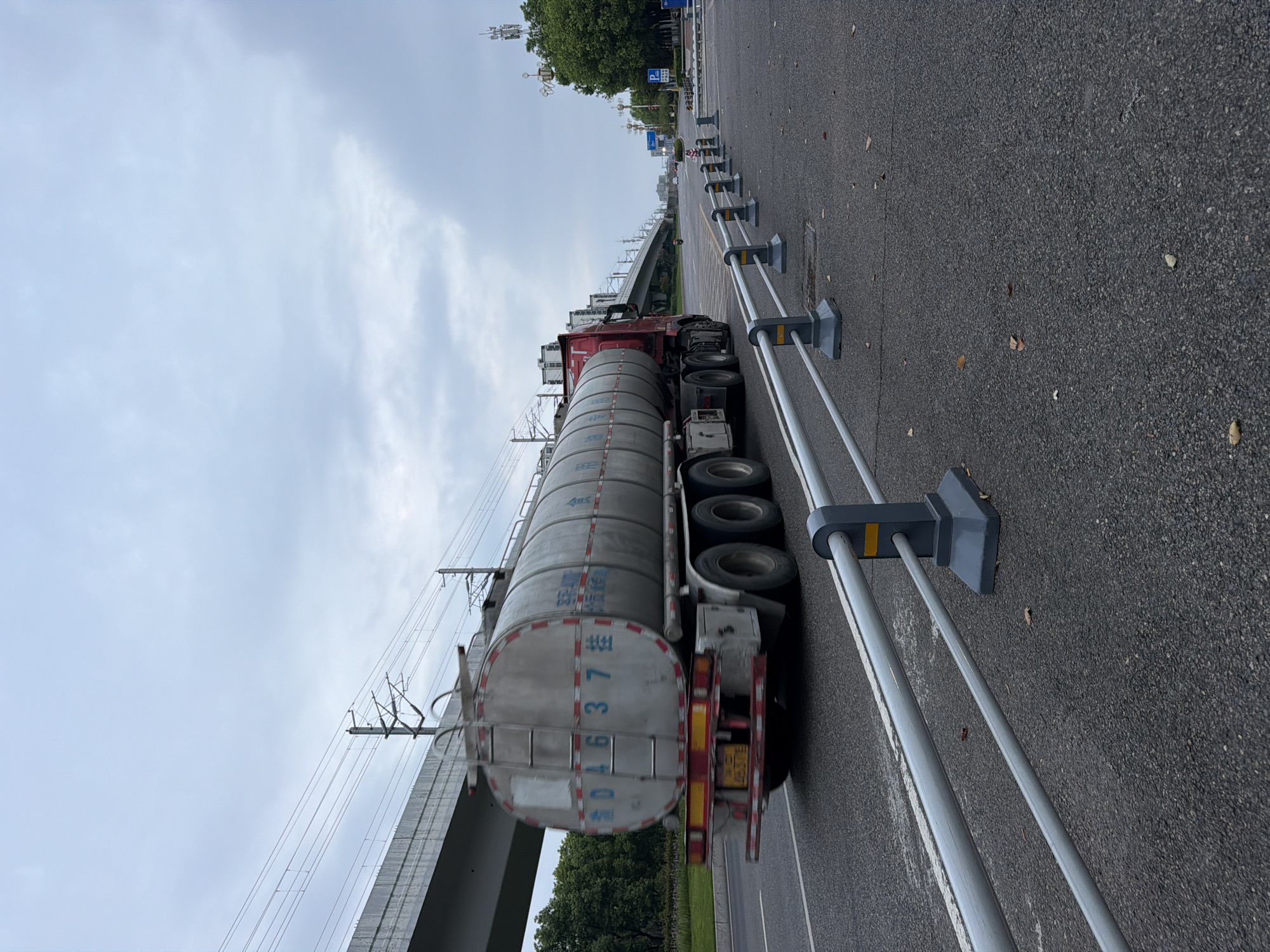 A tanker truck with Chinese characters and license plate "014637" drives on a road in Jiangjin, China, under an overcast sky. The truck is silver with red accents and has multiple axles. The road is asphalt with white lane markings and a metal barrier running along the right side. In the background, an elevated structure, possibly a highway or railway overpass, is visible, along with trees and distant buildings. The scene appears to be in a suburban or industrial area of Jiangjin, with a focus on infrastructure and transportation.