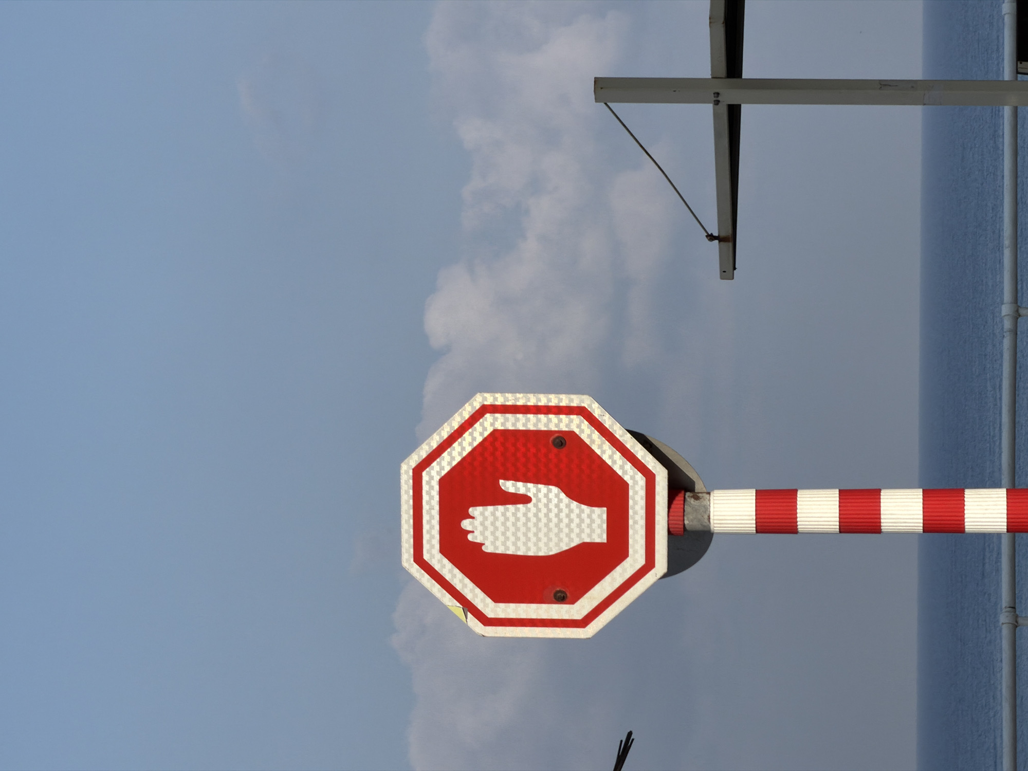 A stop sign with a white hand symbol is mounted on a red and white striped pole, set against a backdrop of a pale blue sky with scattered clouds. The stop sign is octagonal with a red interior and a white border. Two screws are visible on the sign. To the right of the stop sign, a portion of a metal structure with a horizontal beam and a vertical support is visible. In the background, a calm blue body of water, likely the Mediterranean Sea, stretches across the horizon, with a white railing in the foreground. The scene appears to be in Bat Yam, Israel, based on the context provided.