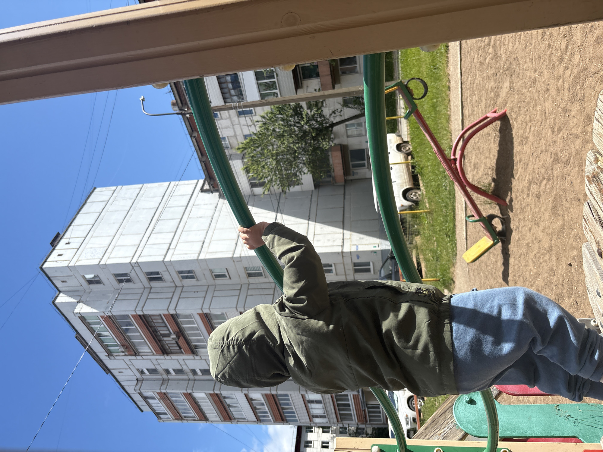A child, viewed from the back, wearing a green hooded jacket and blue pants, stands on a playground structure made of metal bars. The child is reaching up to grasp a horizontal green bar. In the background, a tall, white apartment building with numerous windows and balconies dominates the scene. A tree with green foliage stands to the right of the building. The playground has a sandy surface, and a red and yellow seesaw is visible in the middle ground. White cars are parked in front of the apartment building, and a street lamp is visible in the distance. The sky is clear and blue, indicating a sunny day. This scene appears to be located in a residential area of Chaikovsky, Russia, likely a typical courtyard playground.