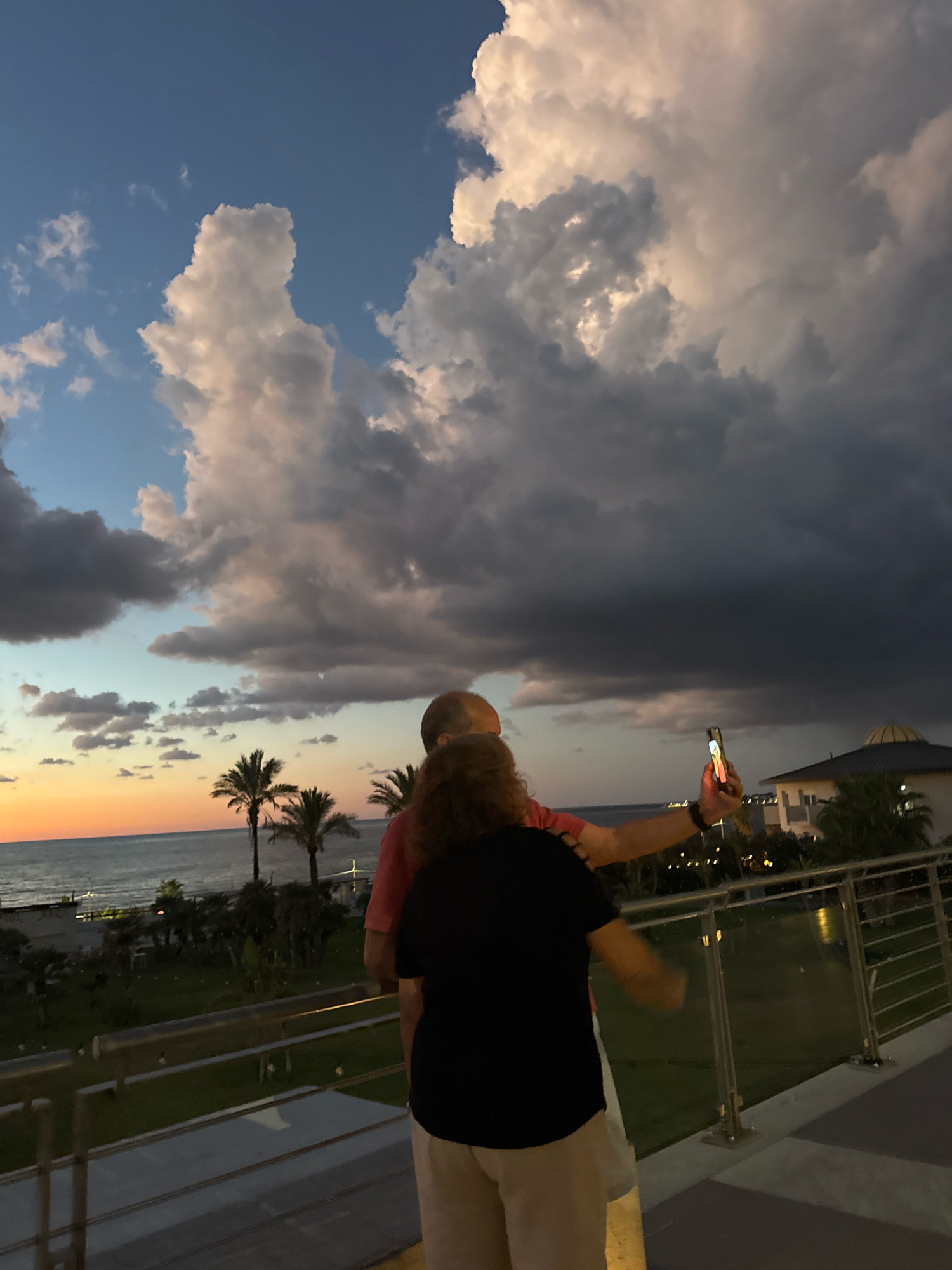 The image captures a scenic view from a balcony or elevated walkway overlooking the sea and coastline at dusk. Two individuals, an older couple, are prominently featured. The male is wearing a pink shirt, and the female is wearing a black t-shirt and light-colored pants. The female is holding up a smartphone, appearing to take a selfie of the couple. The background displays a dramatic sky filled with large, textured clouds, some illuminated by the setting sun, which casts a warm glow on the horizon where the sea meets the sky. Palm trees are visible along the coastline, adding to the tropical or Mediterranean atmosphere. In the distance, there are buildings with domed roofs, suggesting a resort or hotel. The overall scene conveys a moment of shared experience and appreciation of the natural beauty of the location. The location is the city of Isola delle Femmine, Italy.