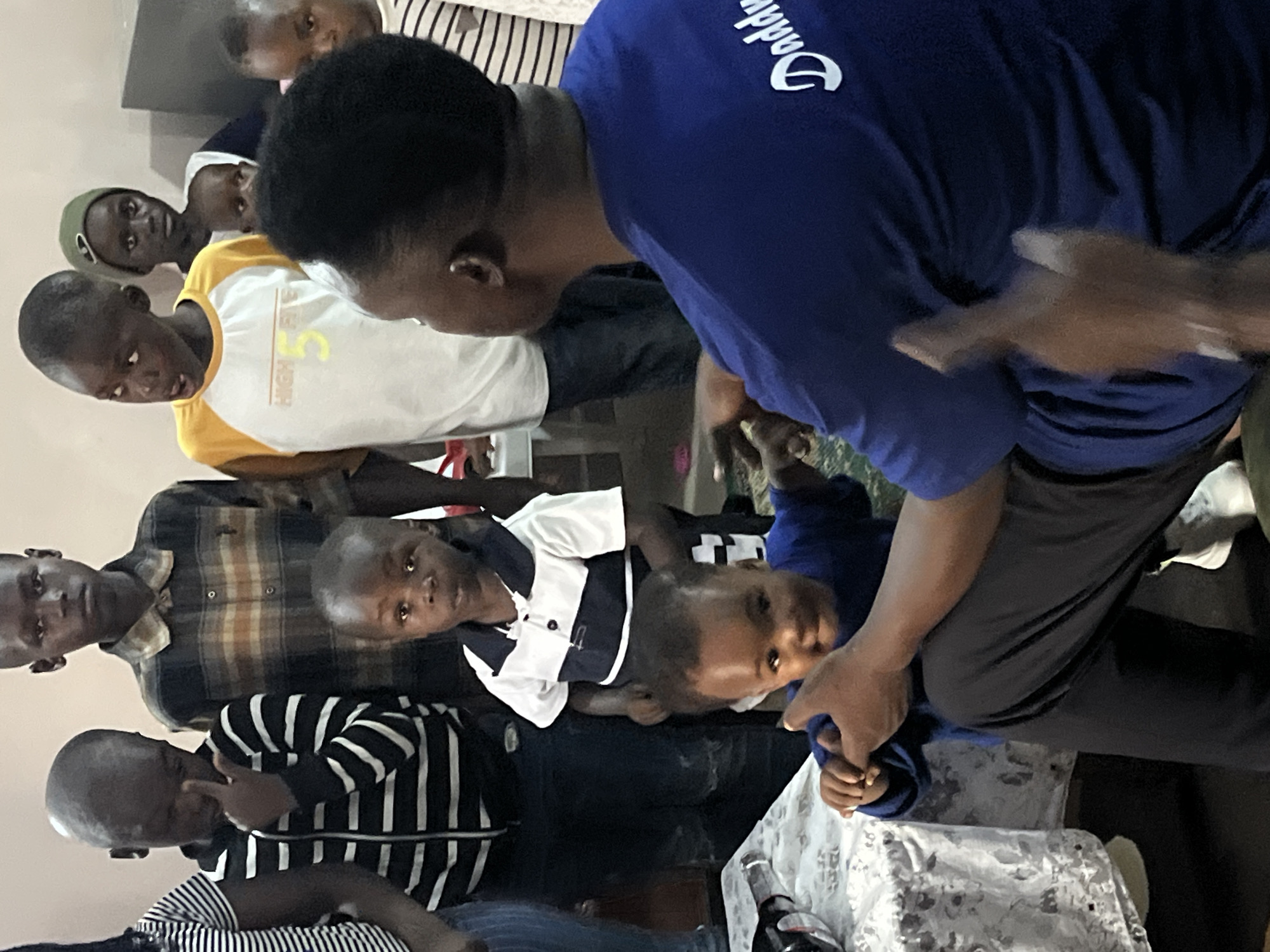The image depicts an indoor scene, likely in Bukuru, Nigeria, with a group of children and an adult. The lighting is moderate, and the background appears to be a plain wall. In the foreground, a young child wearing a blue t-shirt is being held by an adult whose back is to the camera, also wearing a blue t-shirt with white lettering that appears to say "Daddy". The adult's hands are visible, one holding the child's arm and the other raised.
Behind the foreground figures, several children are standing. To the left, a child in a black and white striped jacket is looking down. Next to them, a taller child in a plaid shirt is looking forward. Further back, a child in a yellow and white t-shirt is looking to the left. A child wearing a green beanie is visible behind the child in the yellow t-shirt, and another child in a striped shirt is partially visible on the far right.
To the left of the group, a table covered with a patterned cloth is visible, with a dark object on it that could be a bottle or a container. The floor is dark and appears to be carpeted or tiled. The overall atmosphere suggests a gathering or a casual event.