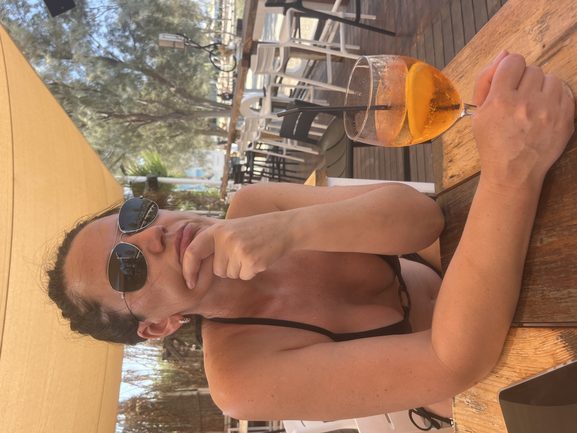 A woman in a black bikini is seated at a wooden table outdoors, resting her chin on her hand and looking to the right with a pensive expression. She is wearing aviator sunglasses and a hoop earring in her visible ear. A glass of orange-colored beverage with a slice of orange and a straw sits on the table in front of her. The background shows a beachside bar or restaurant with tables, chairs, and trees. The overall setting appears to be a sunny day.