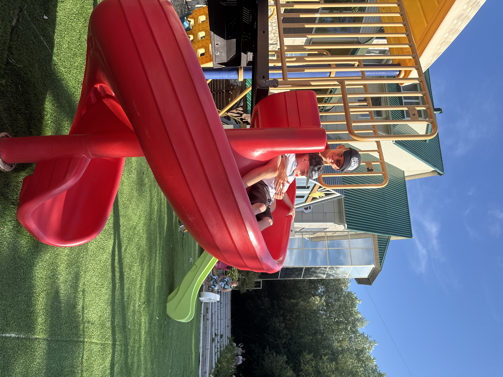 A medium shot captures two young boys on a playground slide in the city of Danilovka (Altai Krai), Russia. The boy in front, wearing a white t-shirt and dark shorts, is seated on the bright red, spiral slide, looking down. Behind him, an older boy in a red t-shirt and a black baseball cap, also looking forward, has his arm around the younger boy. The playground equipment, a mix of red and yellow plastic and yellow metal railings, dominates the foreground. In the background, a building with a green metal roof and large windows is visible, surrounded by trees. The sky is clear and blue, with some wispy clouds. The ground is covered in green artificial turf.
