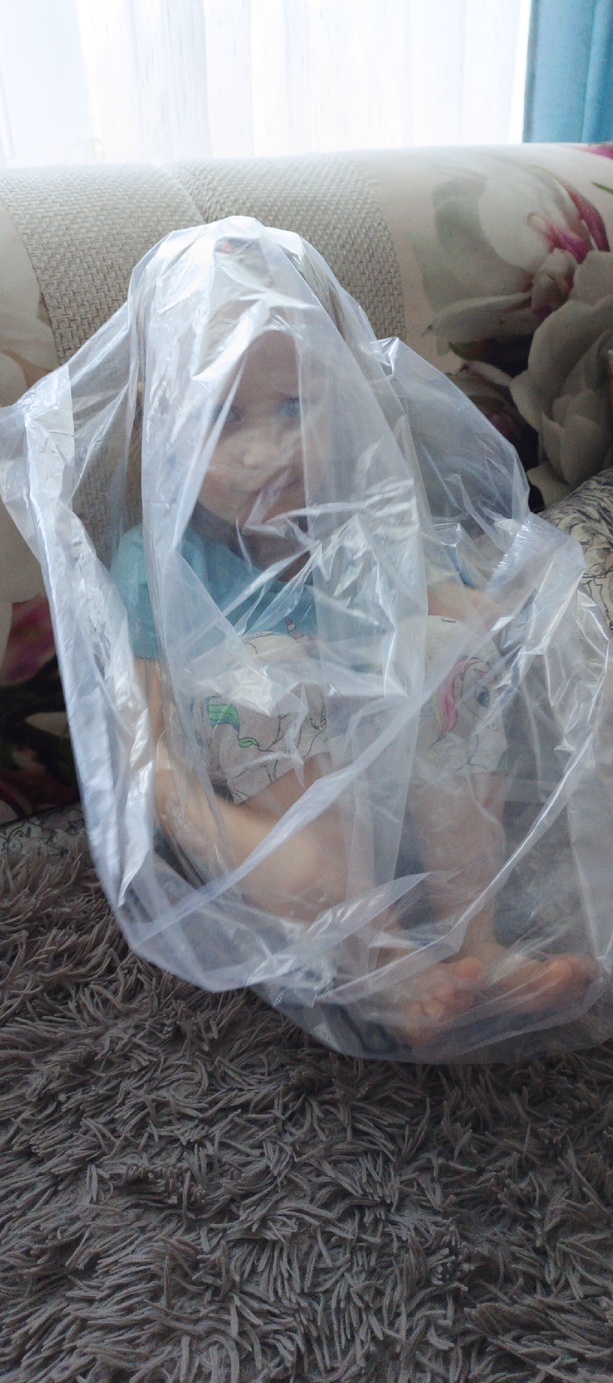 A young child is seated on a floor covered by a shaggy, dark grey-brown carpet. The child's upper body and head are encased within a clear plastic bag, with its face, featuring blue eyes, discernible through the translucent material. The child wears a light blue t-shirt and light-colored shorts with a faint cartoon-like pattern. Behind the child is a sofa, upholstered in a light beige woven fabric, supporting cushions with a white background and prominent floral patterns in shades of pink and green. In the background, a window is visible with sheer white curtains, and a partial blue curtain is present on the right side. The ambient lighting is bright, consistent with daylight. The specific location within the city of Chaikovsky, Russia, is not discernible from the visual information provided.
