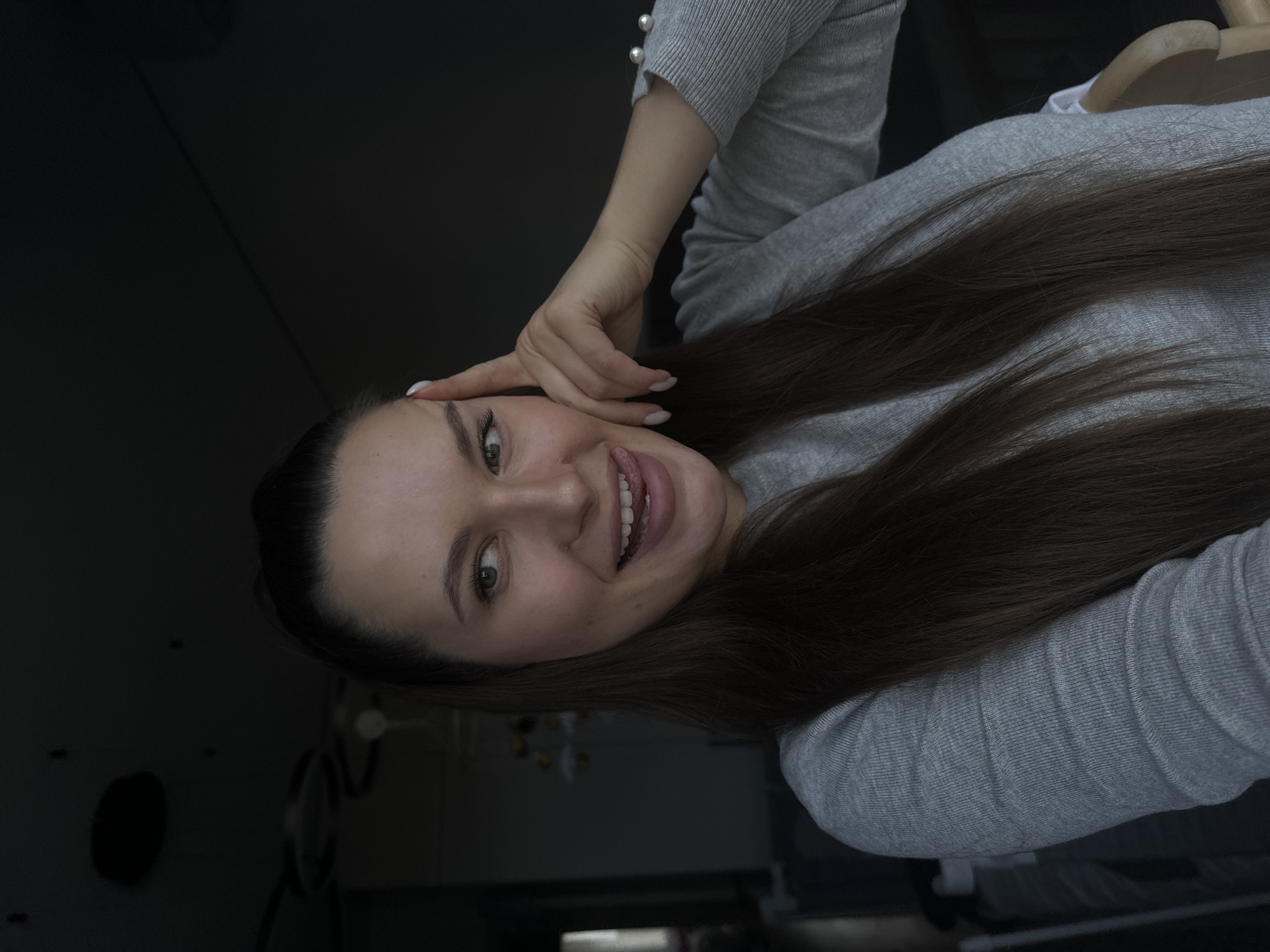 A young woman with long, dark brown hair is captured in a close-up portrait, smiling brightly at the camera. She appears to be standing or sitting upright, with the image rotated 90 degrees counter-clockwise. Her right hand is raised, with her fingers gently touching her temple, indicating a relaxed or playful pose. She wears a light grey, ribbed long-sleeved top, possibly a sweater or sweatshirt, featuring decorative pearl-like buttons on the cuff of her right sleeve. Her facial expression is cheerful, with a wide smile revealing her teeth, and her tongue is slightly visible between her lips, adding to a whimsical or relaxed mood. Her eyes are open and engaging. The background is largely dark and indistinct, suggesting an indoor setting with low lighting or a shallow depth of field blurring the surroundings. No specific objects or discernible details are visible in the background, making it hard to identify the exact location visually, though the provided context places it in Akulovo (Gp Kubinka), Russia. There is no visible text in the image.