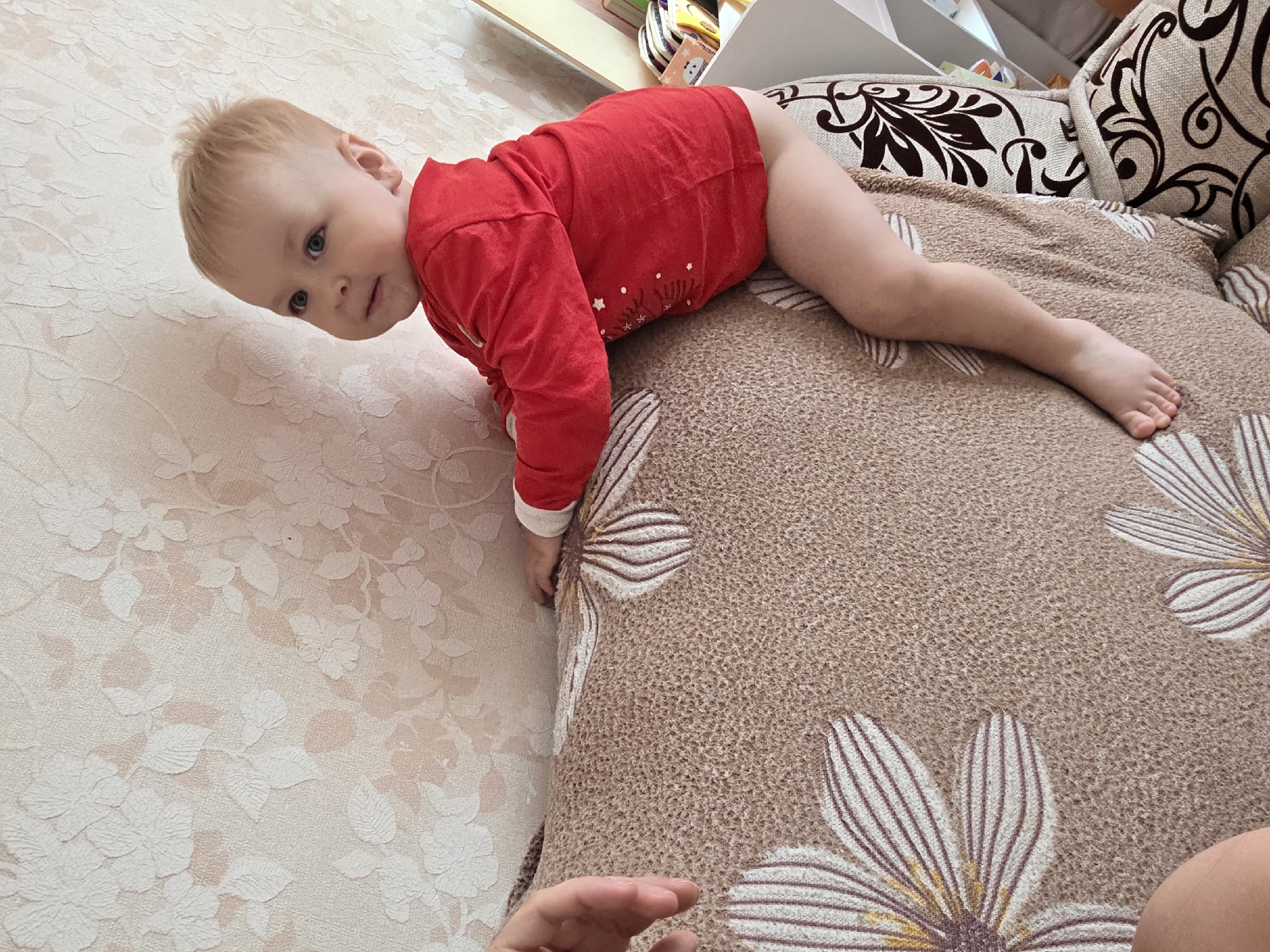A baby in a red long-sleeved onesie is lying on a brown cushion with white floral patterns. The baby's head is turned towards the viewer, and the body is positioned diagonally across the cushion. The background includes a floral-patterned wall and other cushions with dark patterns. A hand is visible at the bottom of the frame.