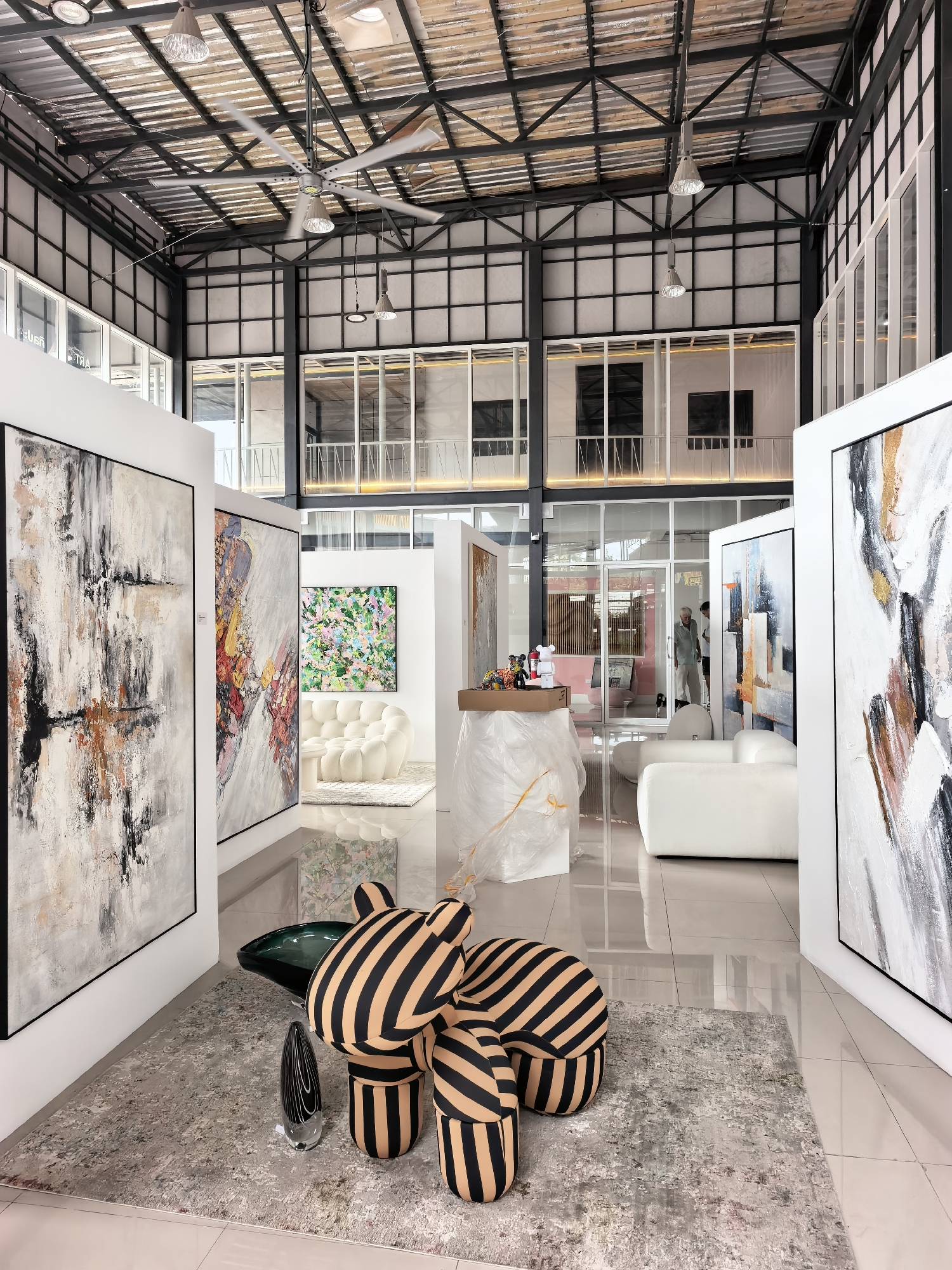 The image depicts the interior of a contemporary art gallery, likely located in a commercial or industrial building with an open-plan design, characterized by a high, exposed metal ceiling with industrial fans and lighting fixtures. Large-format abstract paintings are displayed on white partition walls, with a prominent piece in the foreground on the left. In the center of the space, a unique, striped, biomorphic armchair sits on a textured rug. Adjacent to the armchair is a dark, sculptural glass coffee table and a tall, slender black glass vase. In the background, white modular sofas are arranged, and a vibrant, colorful abstract painting hangs on a wall. Through glass doors and windows, glimpses of other areas within the gallery or an adjacent building are visible, including the silhouettes of individuals. The flooring appears to be polished concrete or tile, reflecting the ambient light. The overall aesthetic suggests a modern, minimalist art exhibition space. The presence of construction-like elements on the ceiling and some areas covered in protective plastic suggest that the gallery may be newly set up or undergoing renovation. The scene is well-lit, creating a bright and airy atmosphere. While the prompt mentions Ko Samui District, Thailand, there are no specific visual cues within the image that definitively confirm this location.