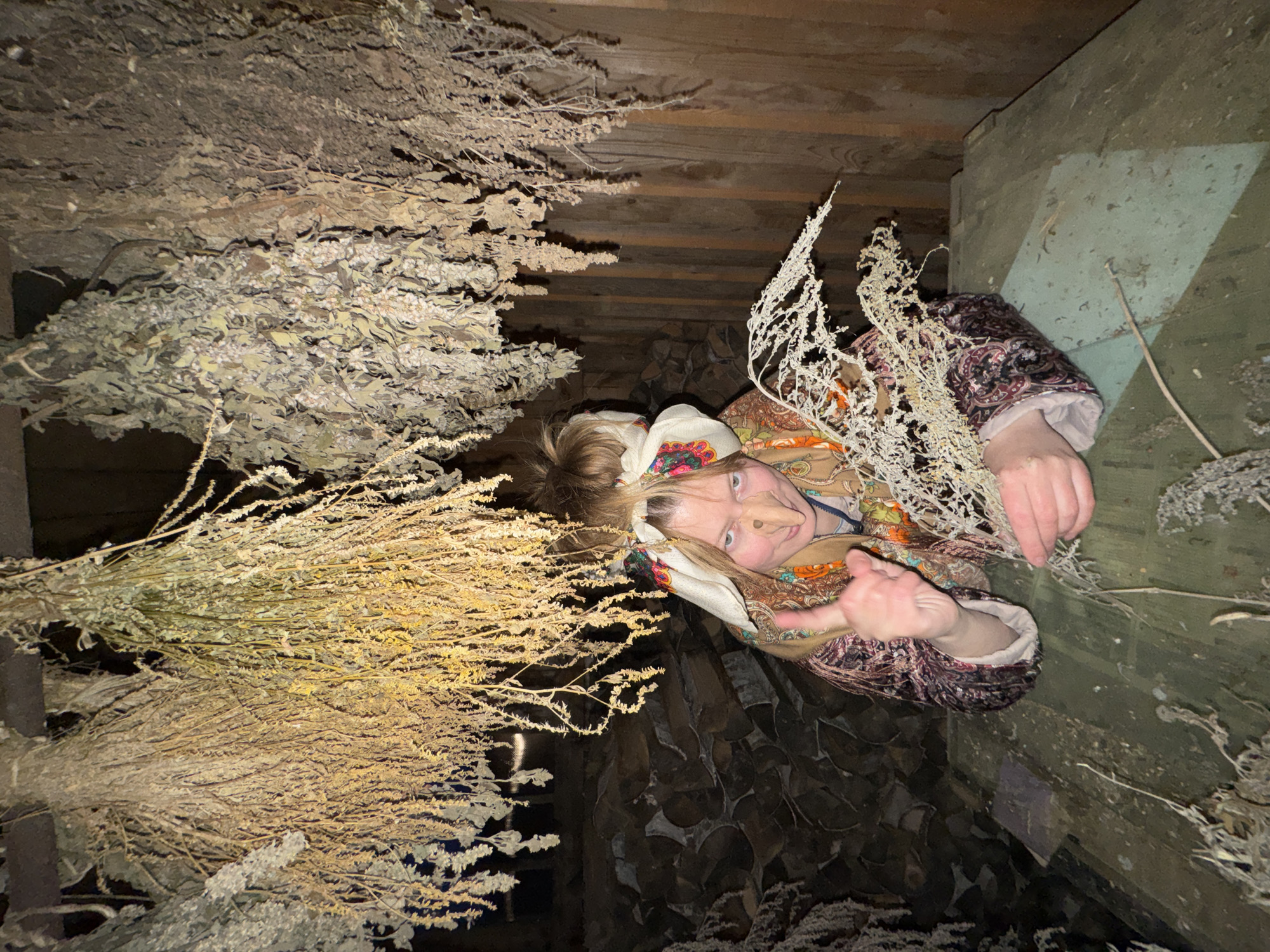 The image appears to be taken indoors. A person with fair skin and blonde hair, wearing a patterned headscarf and a patterned jacket, is visible. The person appears to be in a confined space, surrounded by bundles of dried plants hanging from the ceiling and possibly stacked on the floor. The background includes wooden beams and stacked logs, suggesting a cellar or similar storage area. The location is Вассята, Russia.