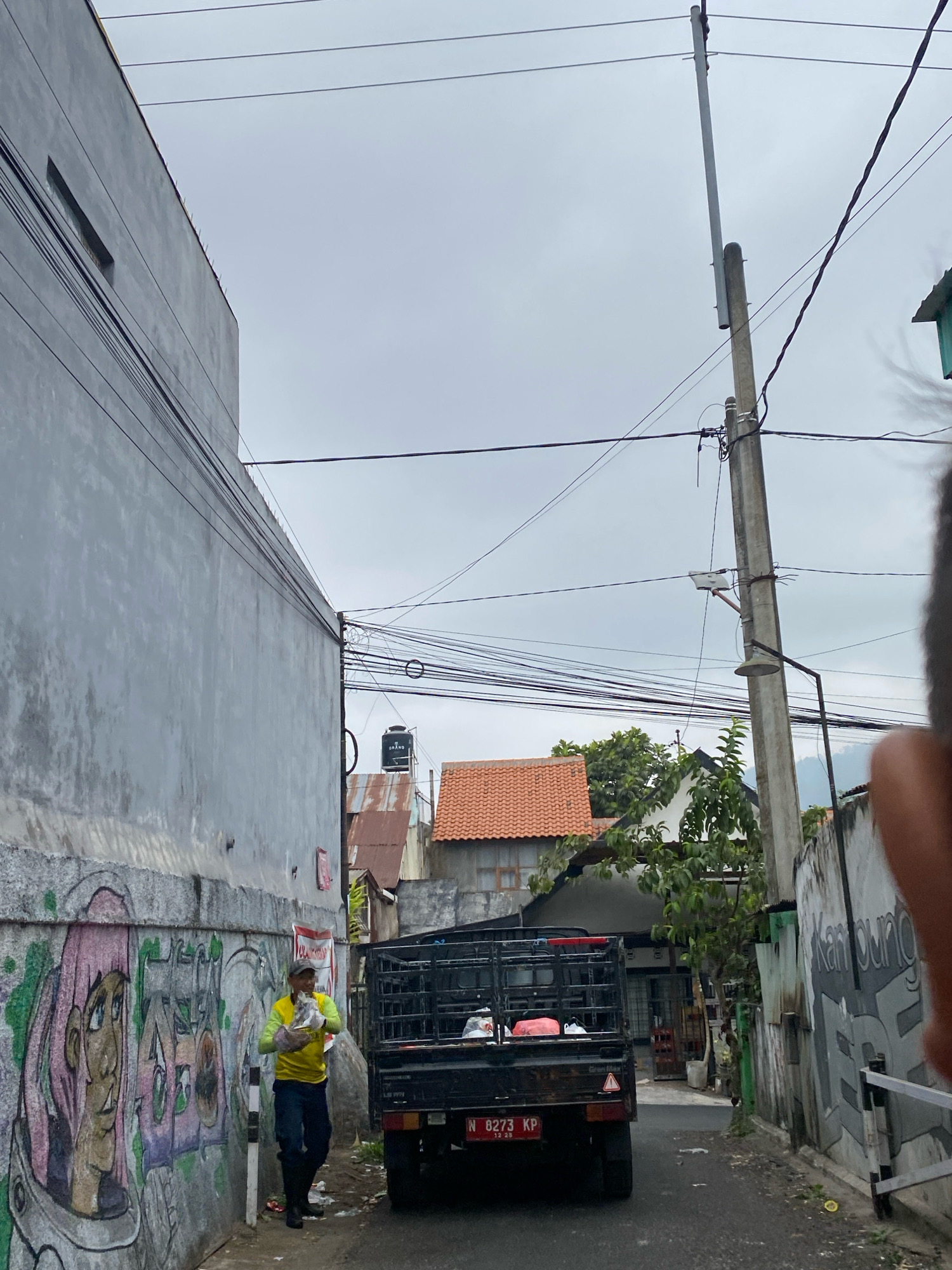An overcast, outdoor shot captures a narrow street lined with buildings and an abundance of electrical wires overhead. On the left, a gray wall is adorned with colorful graffiti depicting a person in a helmet. A man wearing a yellow long-sleeved shirt, blue pants, and a cap walks down the street carrying a plastic bag filled with items. To his right, a black pickup truck is parked. In the background, various buildings with red-tiled roofs are visible, along with a black water tank on a rooftop. A concrete utility pole stands near the center of the image, supporting numerous wires and a street lamp. The ground is paved, and debris is scattered along the sides of the street.